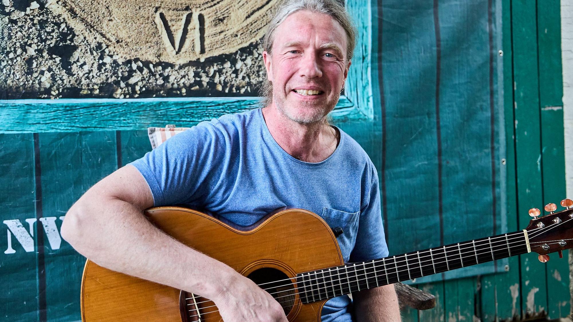 Oliver Kerk hat seine Gitarre in der Hand und sitzt vor einer Holzwand.