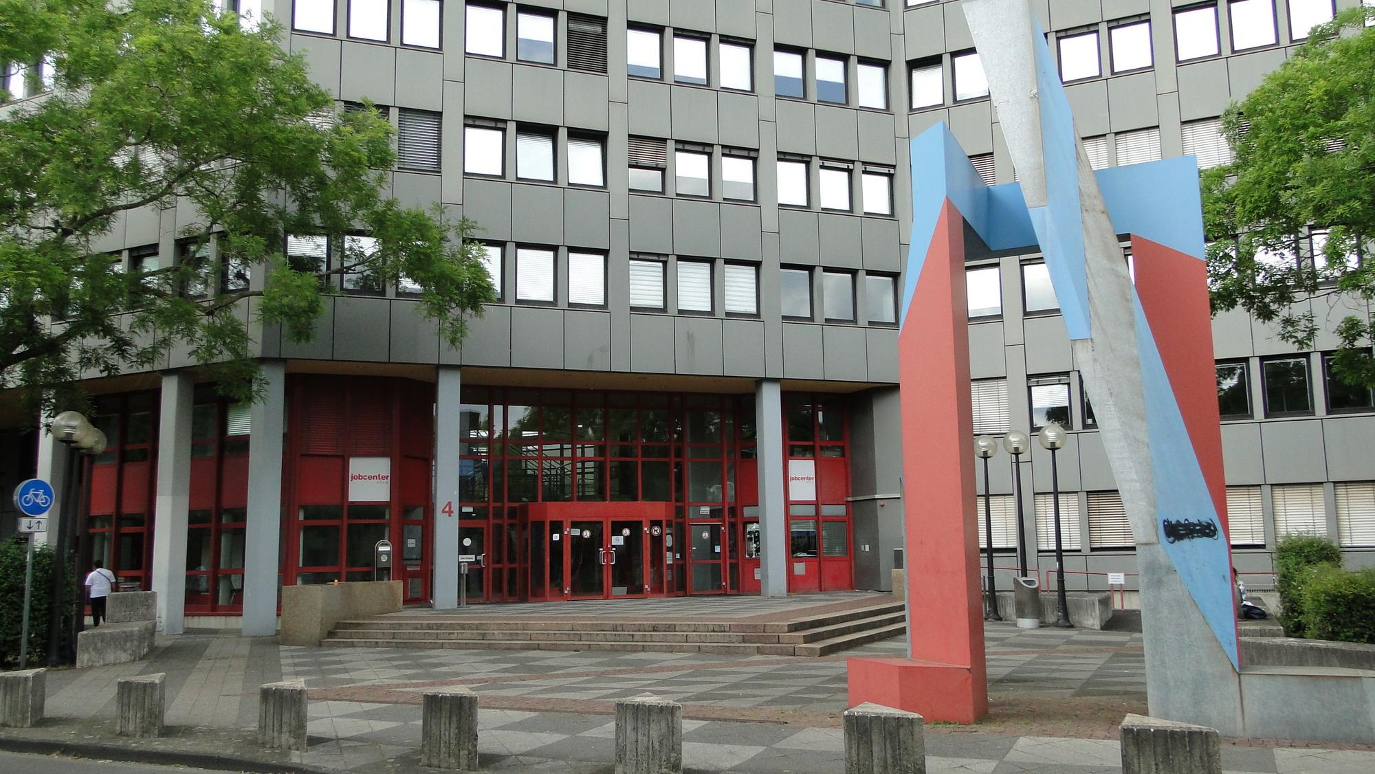 Die Zweigstelle des Jobcenters Chorweiler, ein graues mehrgeschossiges Gebäude mit rotem Eingangsbereich.