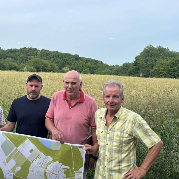 Auf dem Bild ist eine Gruppe von drei Männern und einer Frau zu sehen, die vor einem Feld stehen. Sie halten einen Plan in der Hand.