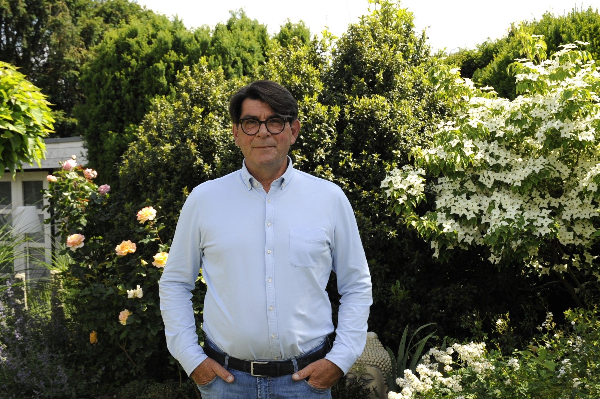 Ein Mann mit dunklen Haaren und hellblauem Hemd steht in einem Garten.