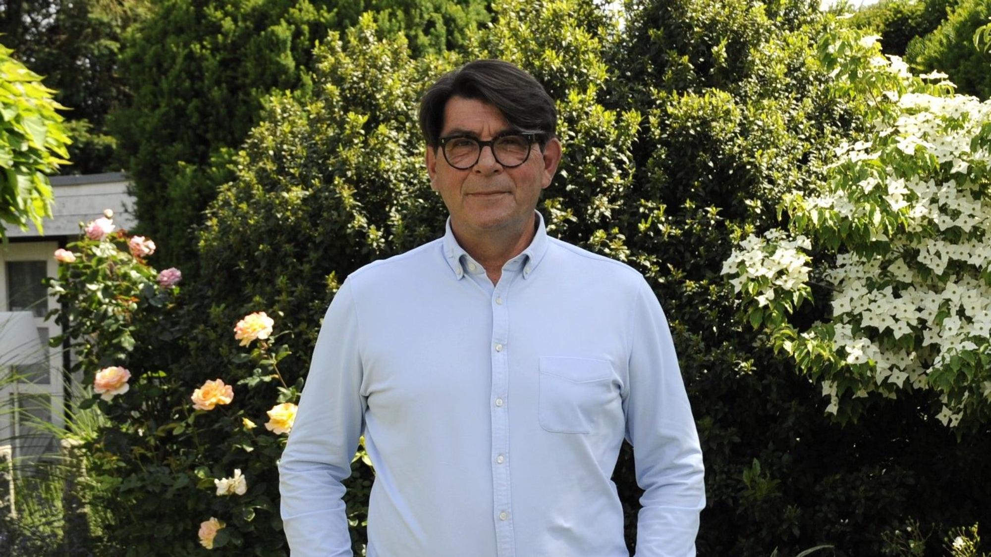 Ein Mann mit dunklen Haaren und hellblauem Hemd steht in einem Garten.