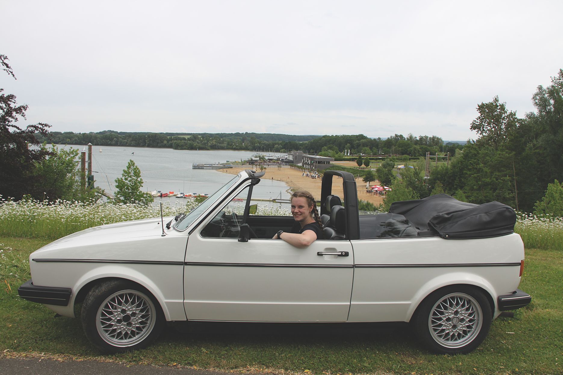 In einem weißen Golf-Cabriolet sitzt eine junge Frau. Im Hintergrund ist der Zülpicher Wassersportsee zu sehen.