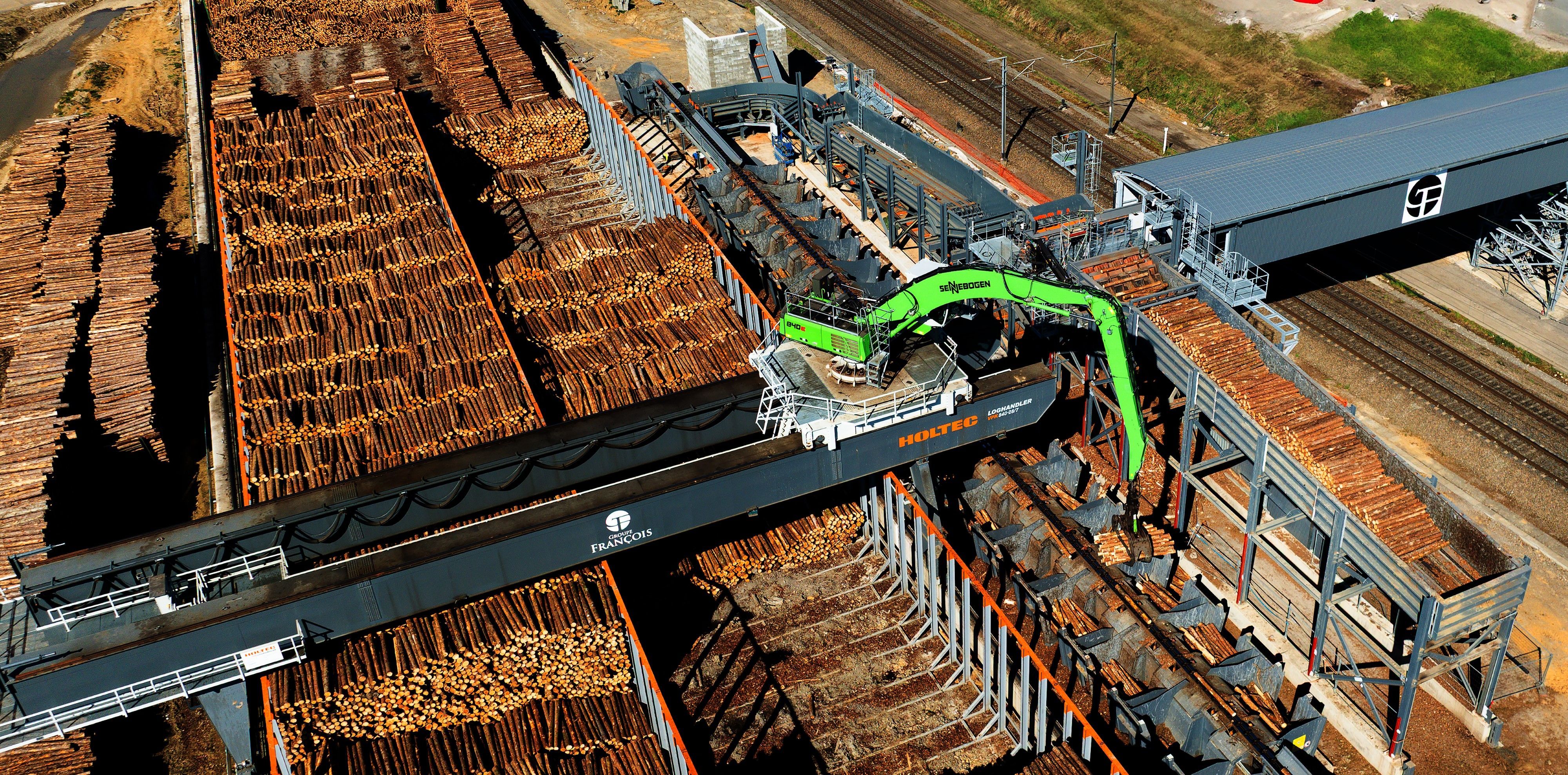 Ein Portalkran bewegt sich in einem Schienensystem mit Brücke über den Holzplatz.