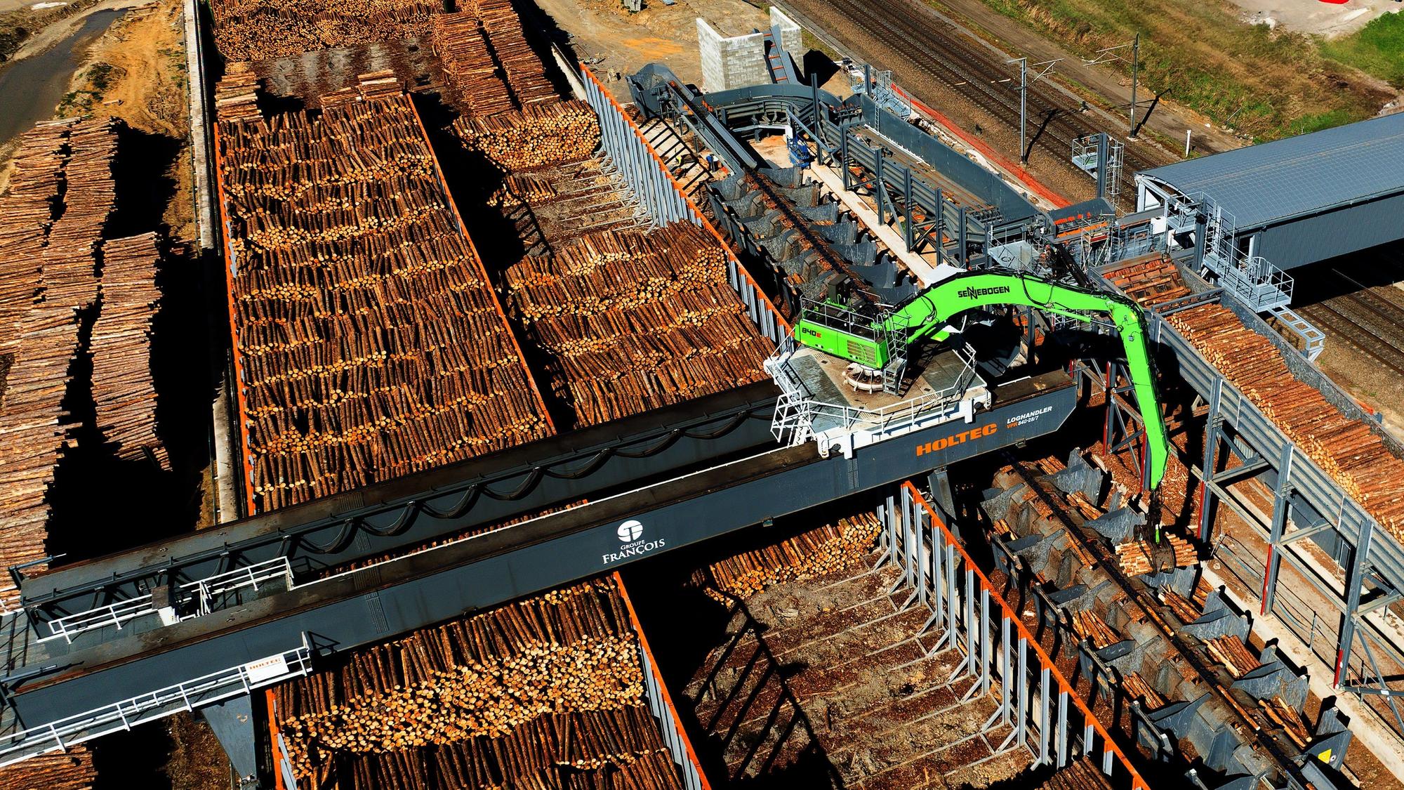 Ein Portalkran bewegt sich in einem Schienensystem mit Brücke über den Holzplatz.