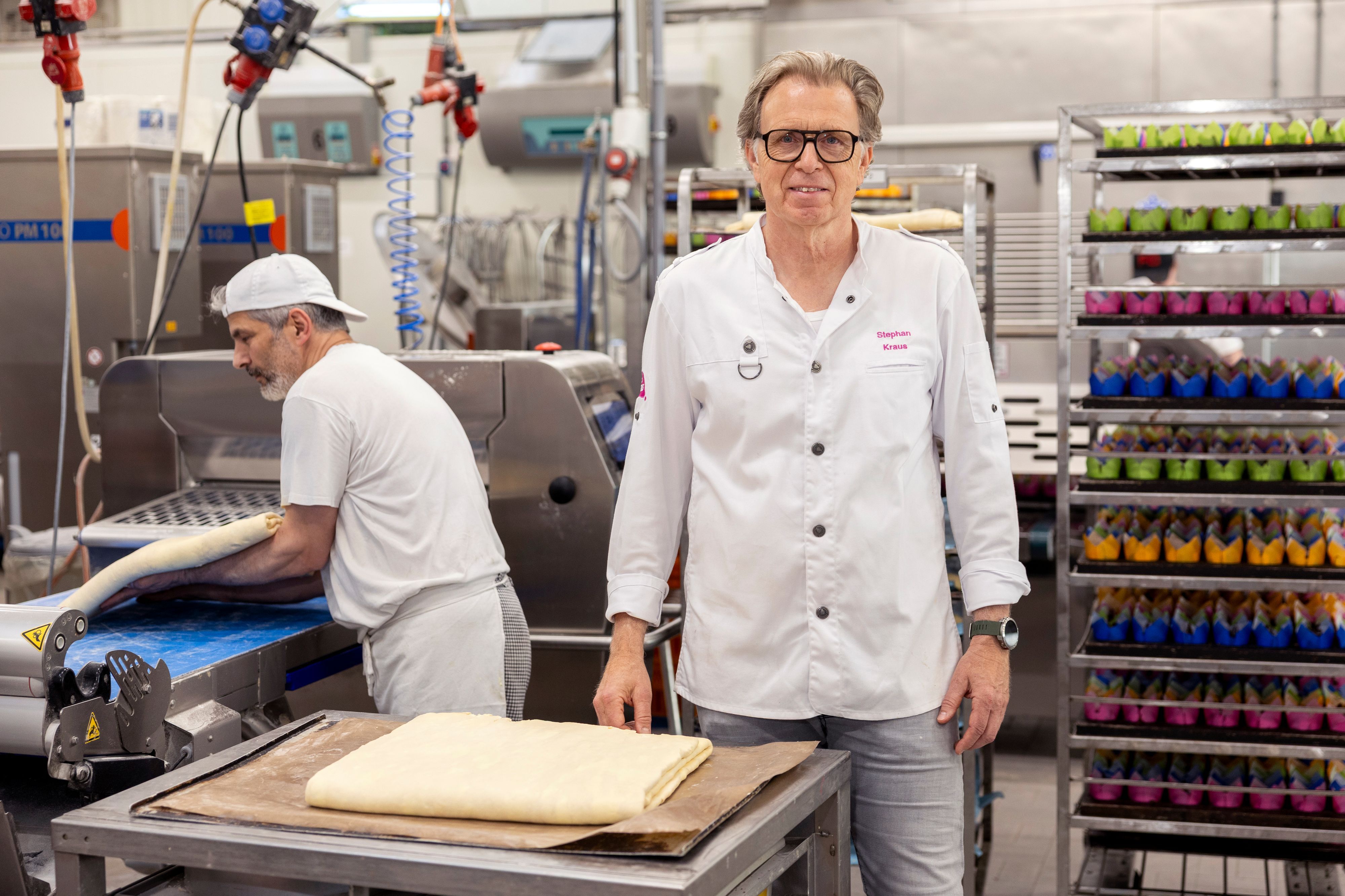 Stephan Kraus steht in der Produktionshalle der Bäckerei Kraus. Hinter ihm hievt ein Mitarbeiter Teig auf ein Band.