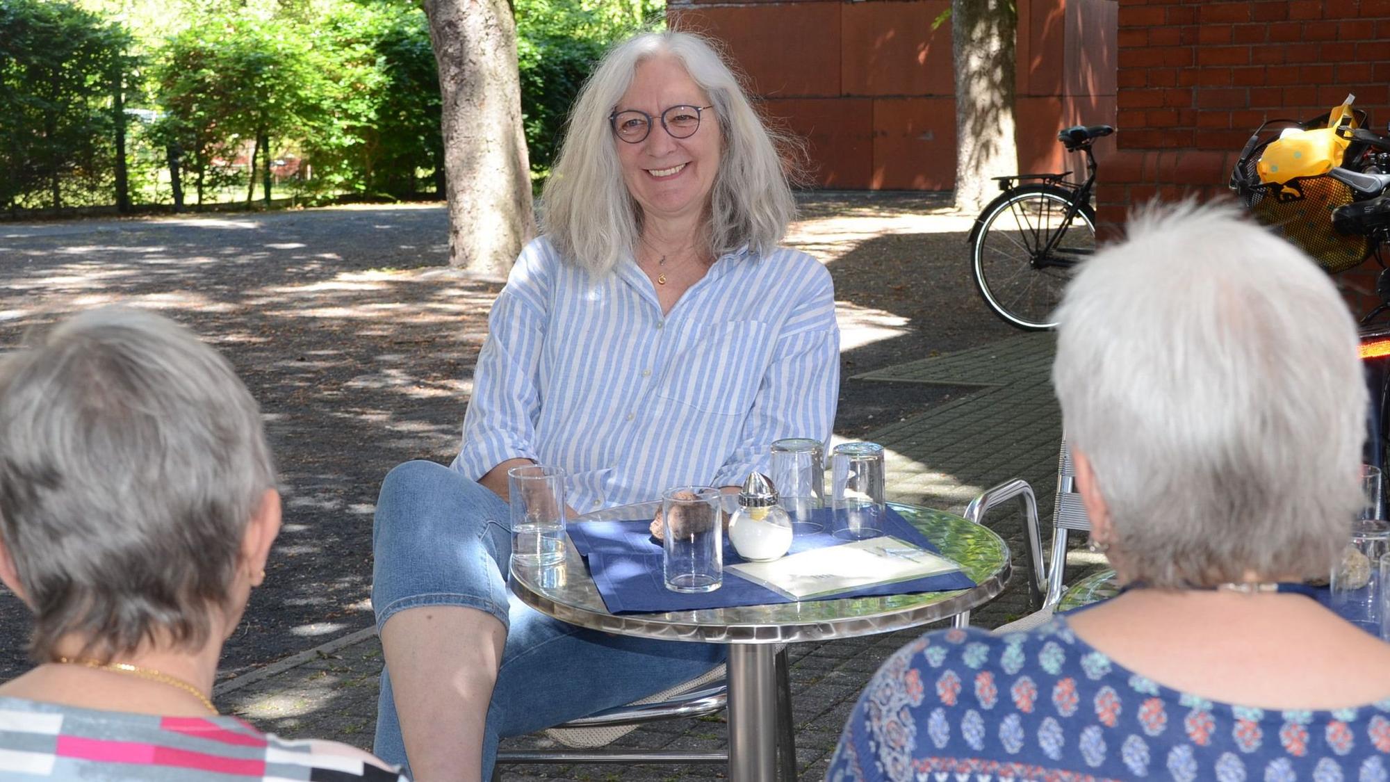 Eine Frau mit langen grauen Haaren sitzt an einem kleinen Tisch unter Bäumen.