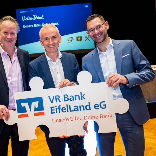 Die drei Bankvorstände halten gemeinsam ein Schild mit dem Logo der Volksbanken, dem Namen „VR Bank EifelLand eG“ und dem Slogan „Unsere Eifel. Deine Bank.““