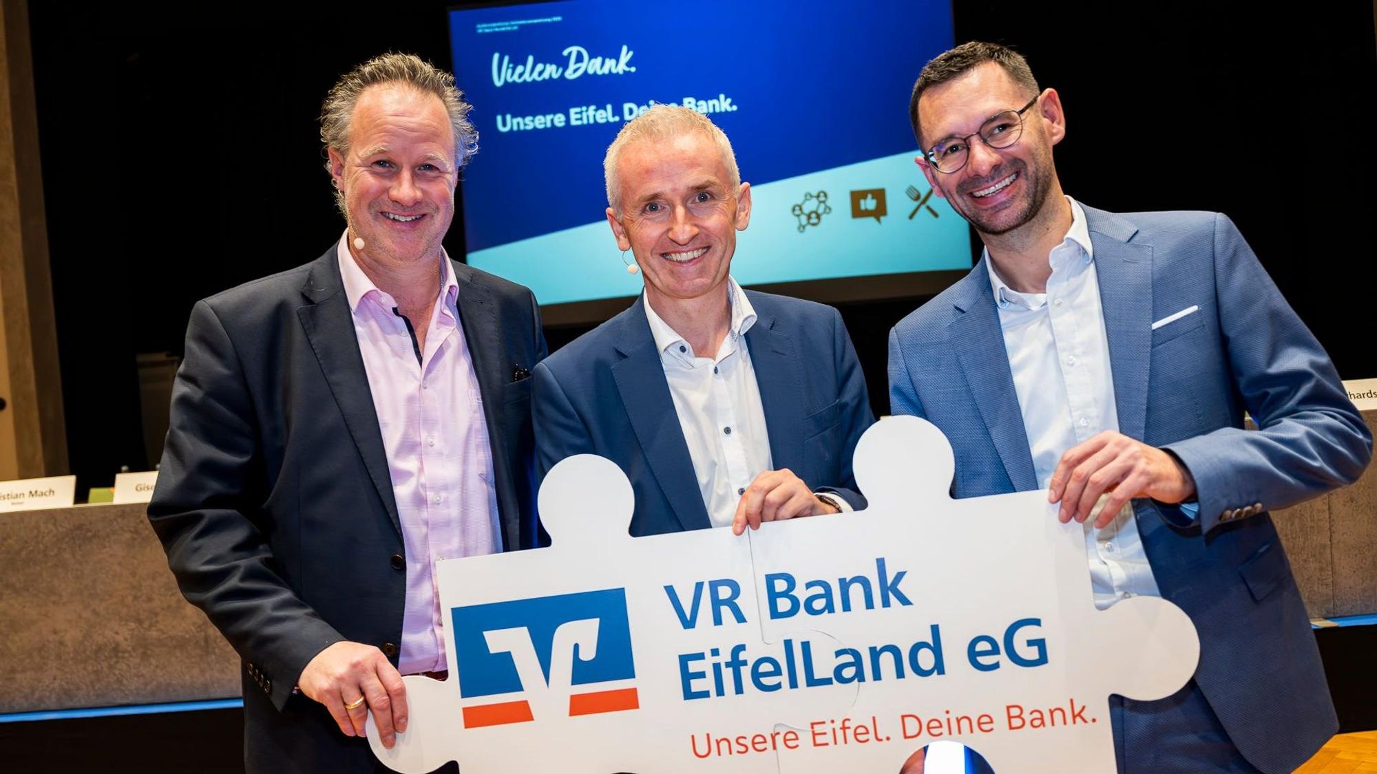 Die drei Bankvorstände halten gemeinsam ein Schild mit dem Logo der Volksbanken, dem Namen „VR Bank EifelLand eG“ und dem Slogan „Unsere Eifel. Deine Bank.““