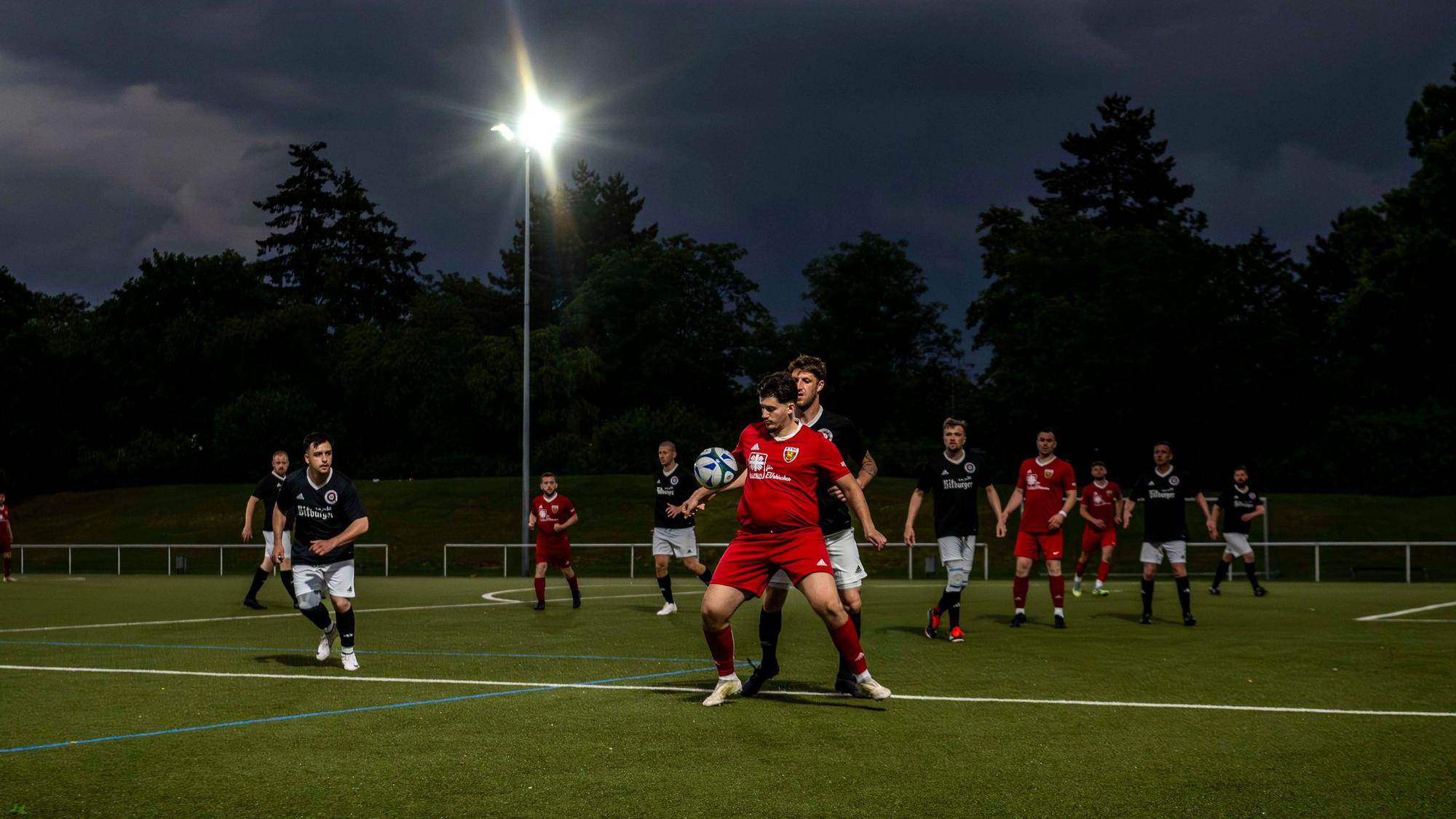 Gewitterwolken tauchen einen Platz in ein dunkles Licht, ein Fußballer in rotem Trikot nimmt einen Ball mit der Brust an.