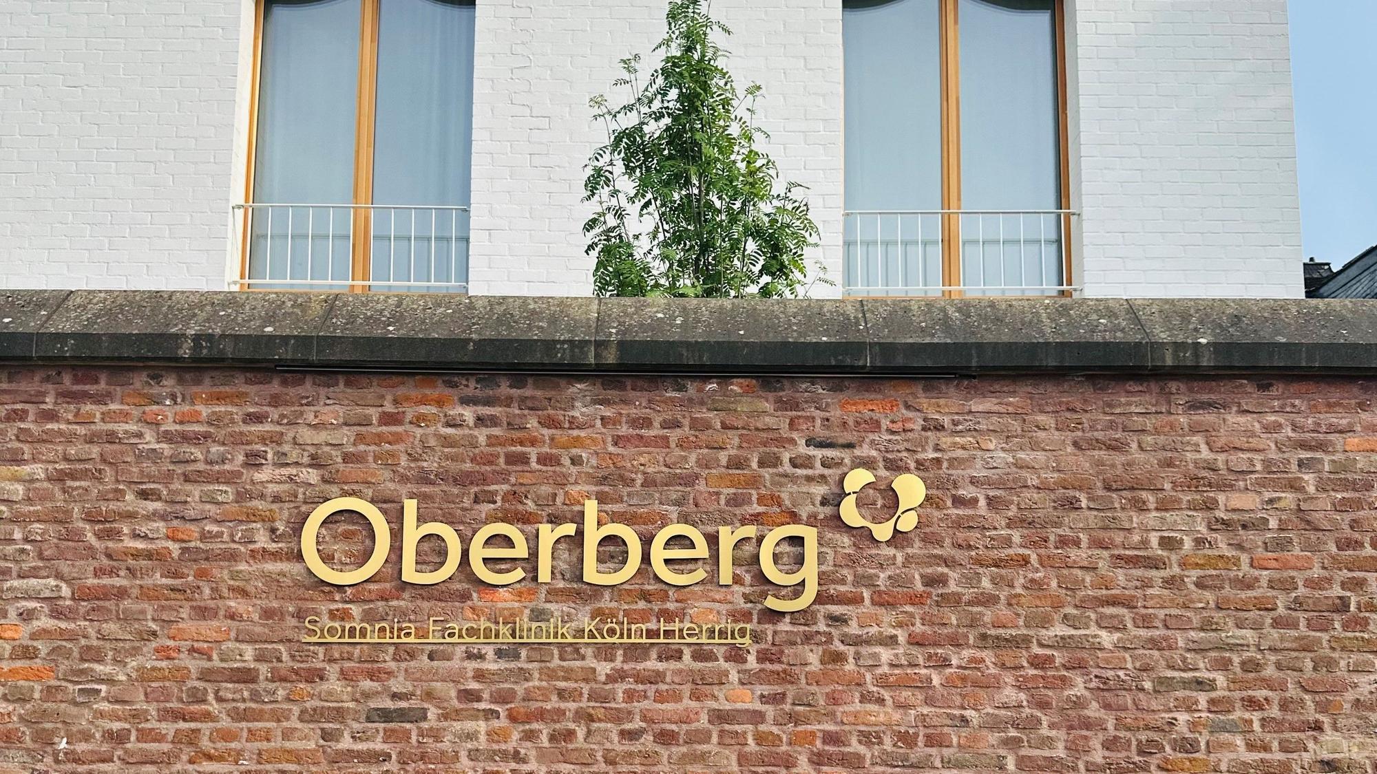 Zu sehen ist eine Mauer vor einem Klinikgebäude mit einem goldenen Schriftzug „Oberberg“.