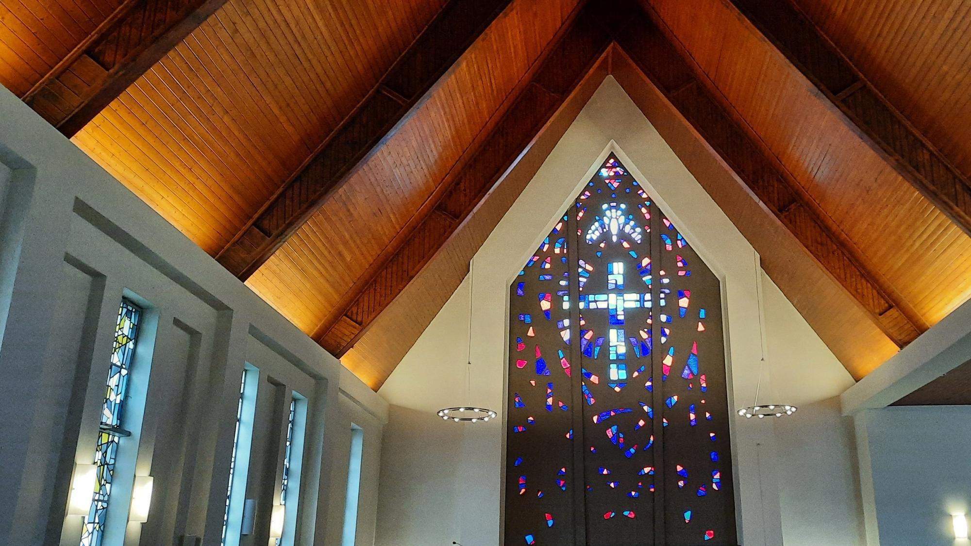 Das Bild zeigt den Innenraum der Kirche. Vorne ist ein Kreuz als buntes Fenstermosaik zu sehen.