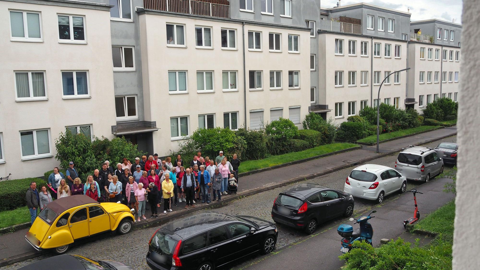 Autos parken an einer Straße, eine Menschengruppe steht auf einer Seite auf dem Bürgersteig.
