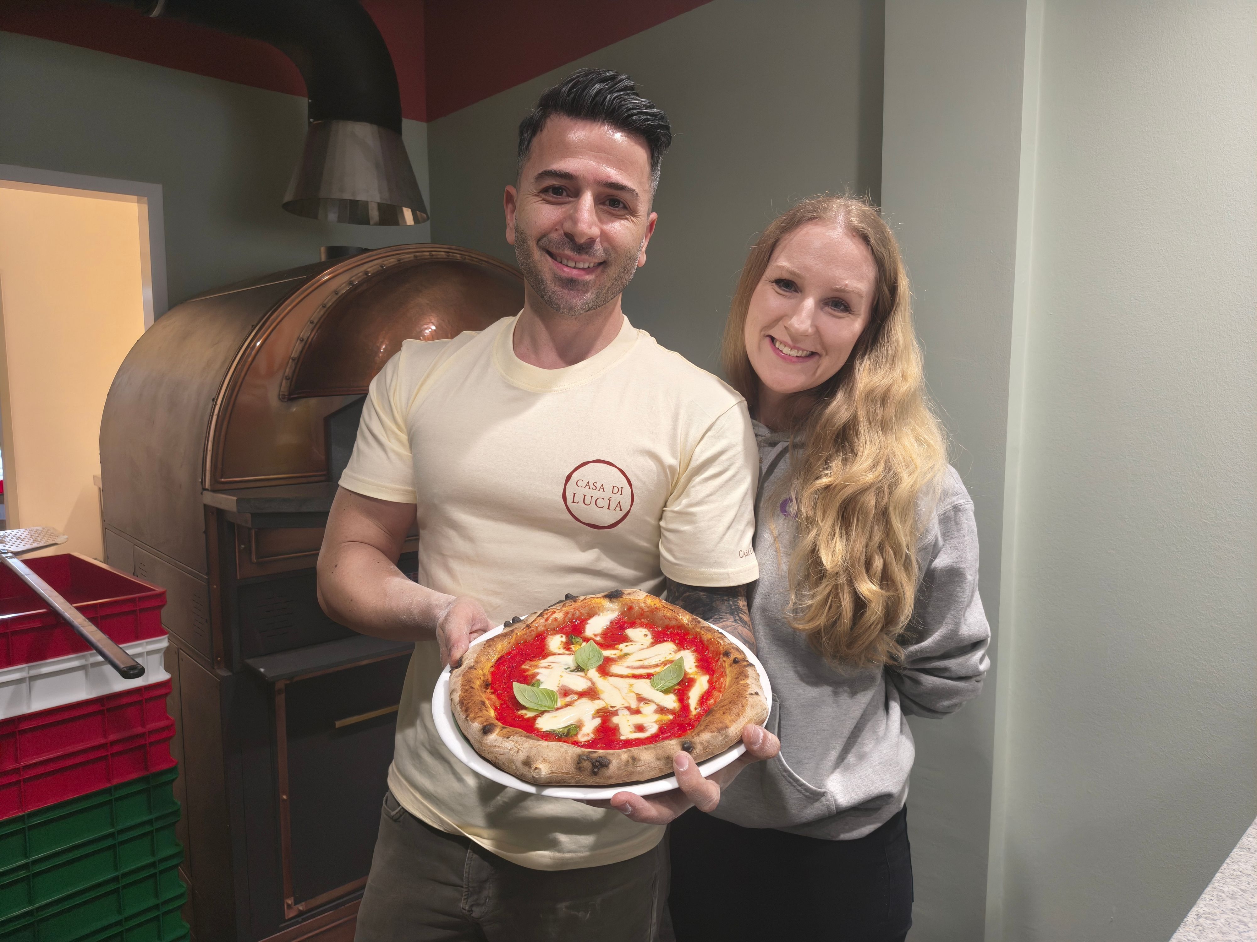 Sinan Batur steht dicht neben seiner Ehefrau Alruna Batur vor einem Ofen und präsentiert eine neapolitanische Pizza auf einem Teller.