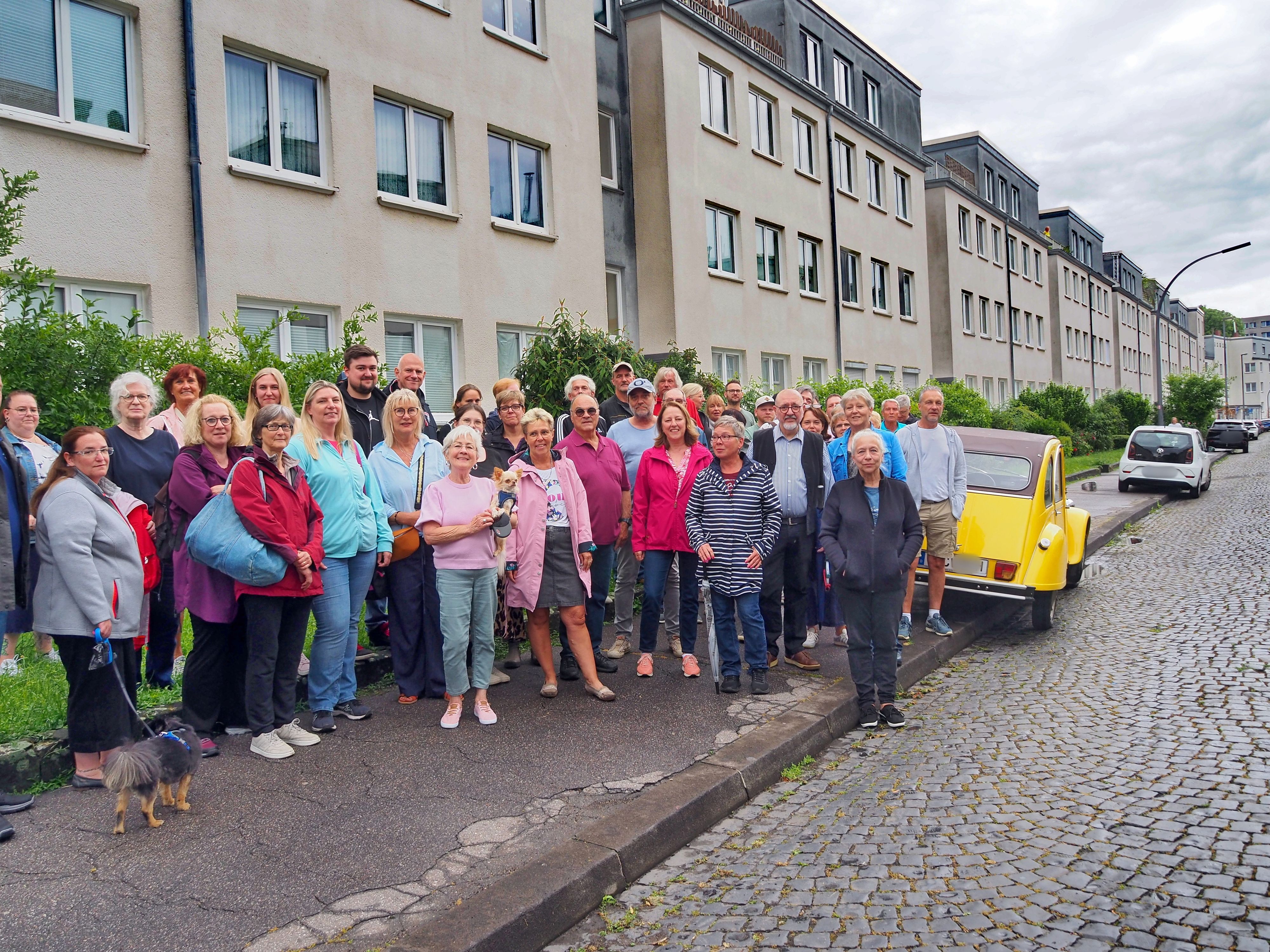 Eine Gruppe Menschen steht auf einem Gehweg, eine Ente parkt auf dem Bürgersteig.