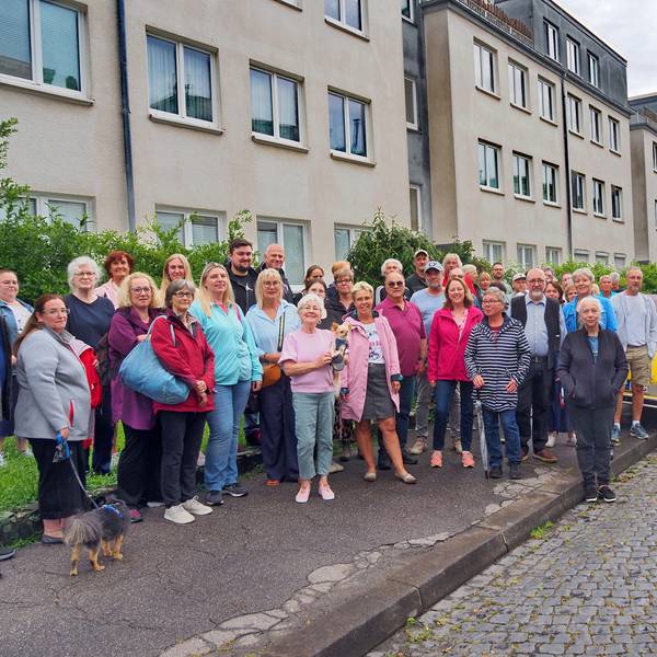 Eine Gruppe Menschen steht auf einem Gehweg, eine Ente parkt auf dem Bürgersteig.