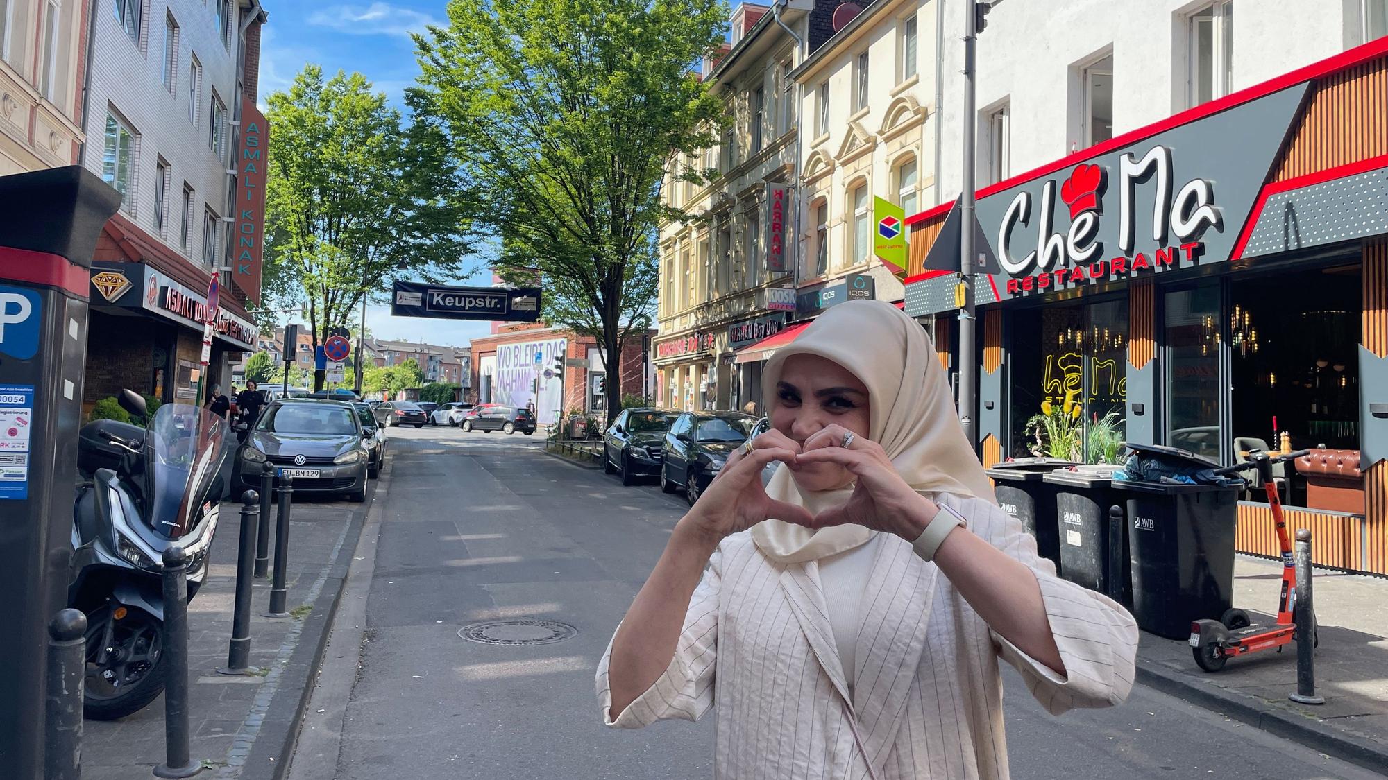 Eine Frau mit hellem Gewand und Kopftuch steht auf einer schmalen Straße und formt mit den Händen ein Herz.