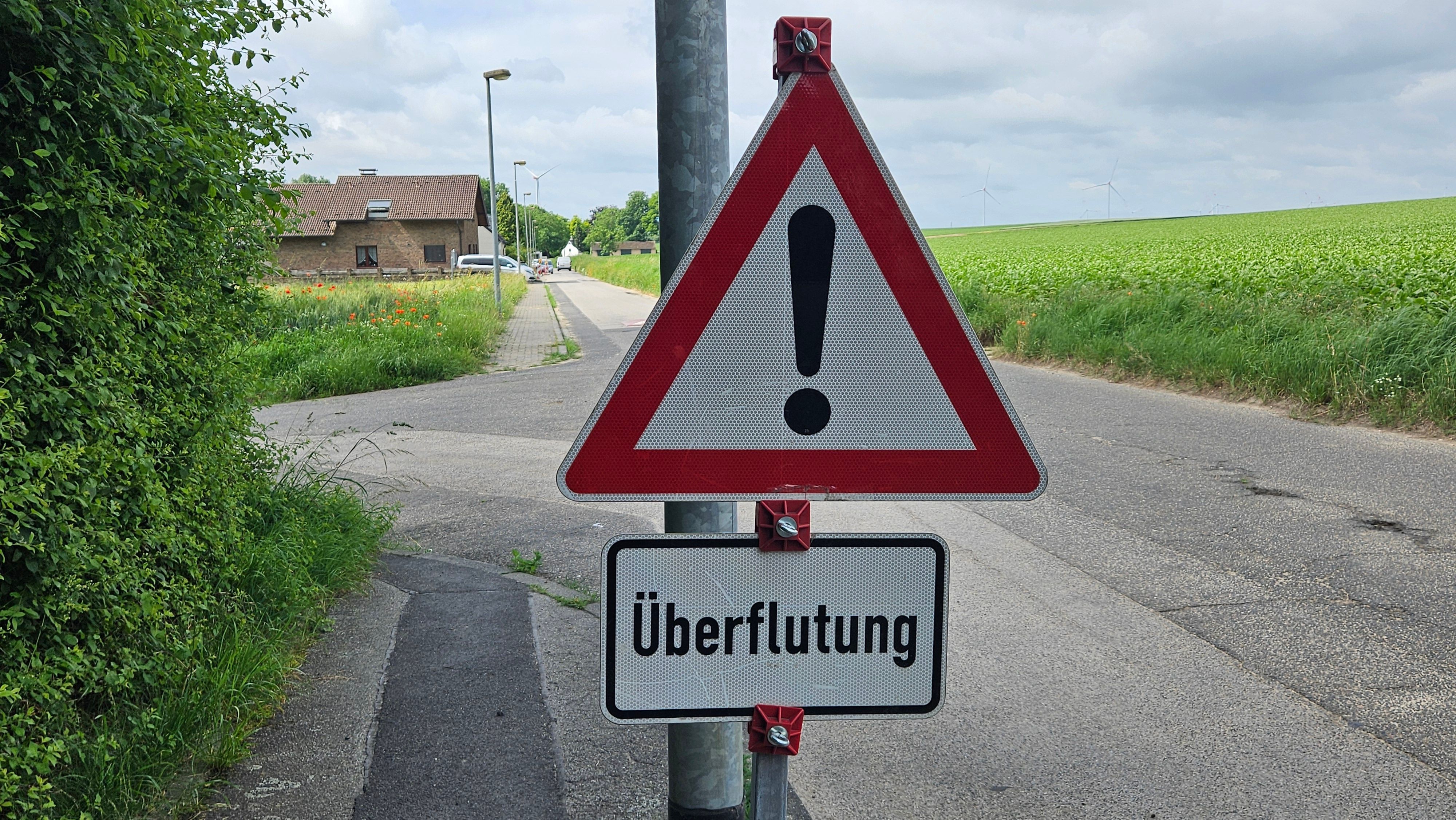 Zu sehen ist ein Warnschild mit der Aufschrift „Überflutung“, im Hintergrund ein Haus und ein Acker.