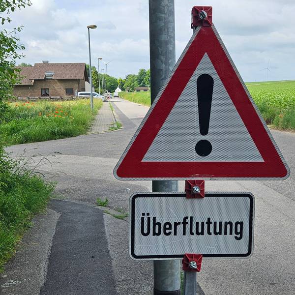 Zu sehen ist ein Warnschild mit der Aufschrift „Überflutung“, im Hintergrund ein Haus und ein Acker.