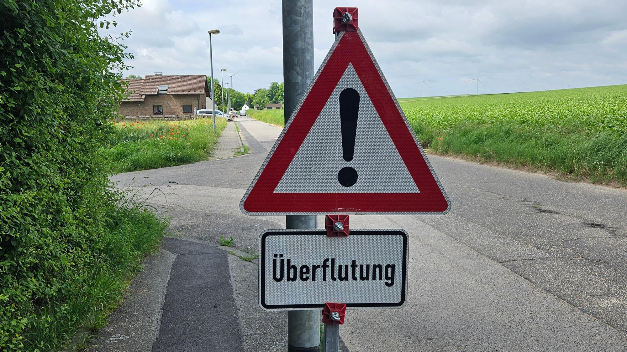 Zu sehen ist ein Warnschild mit der Aufschrift „Überflutung“, im Hintergrund ein Haus und ein Acker.