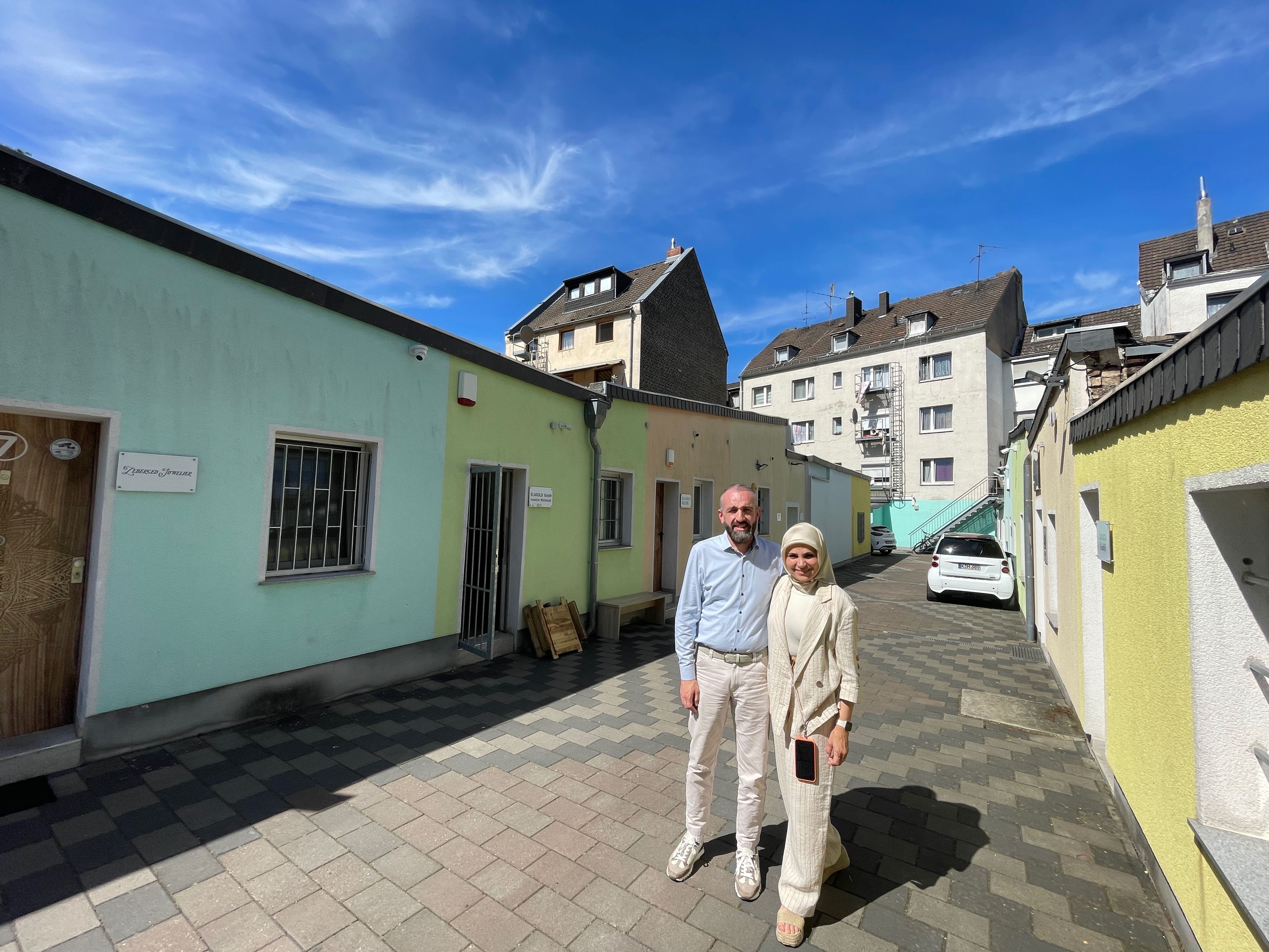 Ein Mann mit hellblauem Hemd und heller Hose steht neben einer Frau in hellen Sachen und mit hellem Kopftuch.