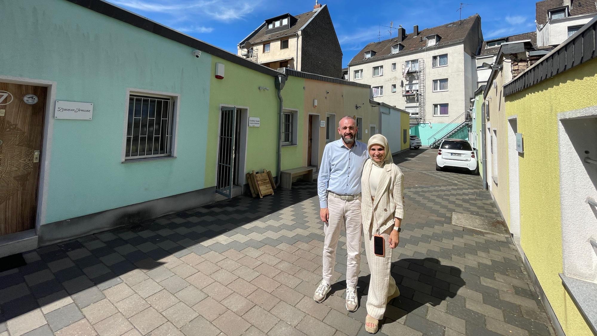 Ein Mann mit hellblauem Hemd und heller Hose steht neben einer Frau in hellen Sachen und mit hellem Kopftuch.