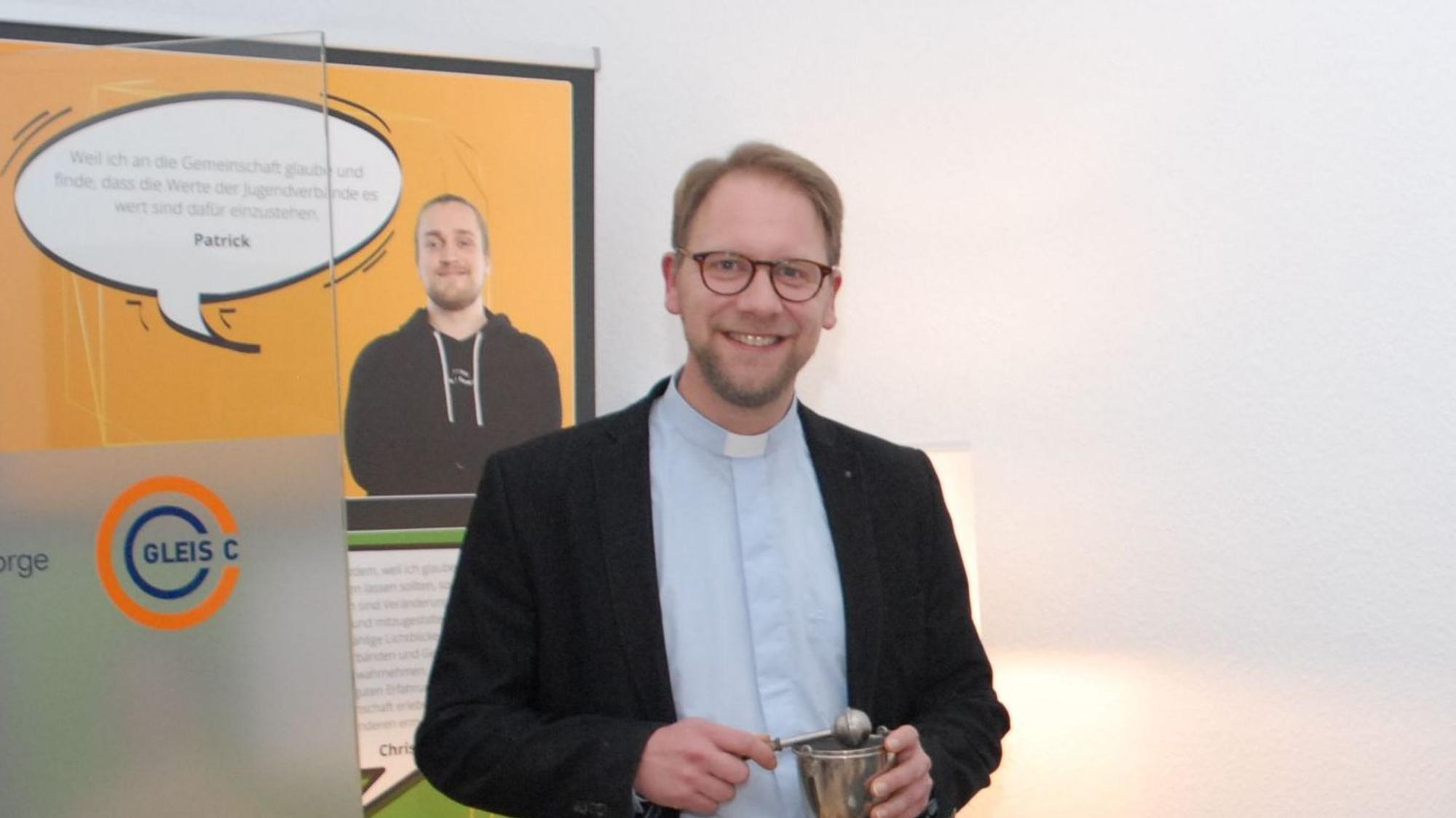 Tobias Schwaderlapp hält die Utensilien für eine Segnung in Händen, er steht vor einem Plakat.