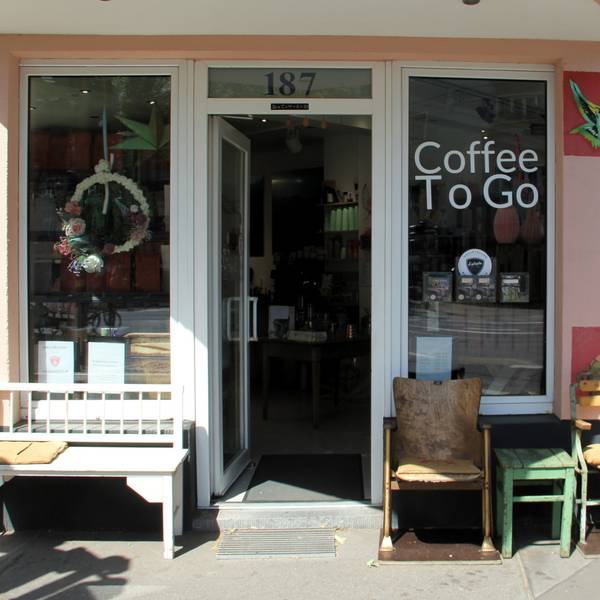 Die Front eines Cafés ist zu sehen. Es stehen Bänke und Stühle davor, an der Seite ist der Schriftzug Urwaldkaffee angebracht.