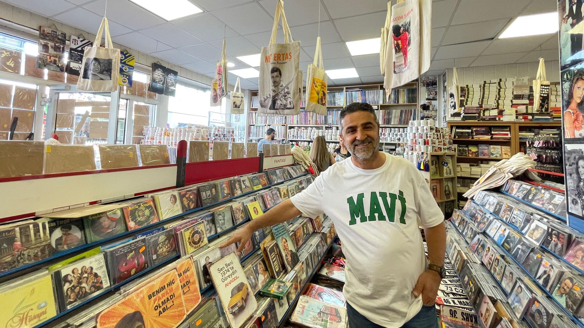 Ein Mann mit dunklem Haar und Dreitagebart steht zwischen Regalen voller Schallplatten und CDs.