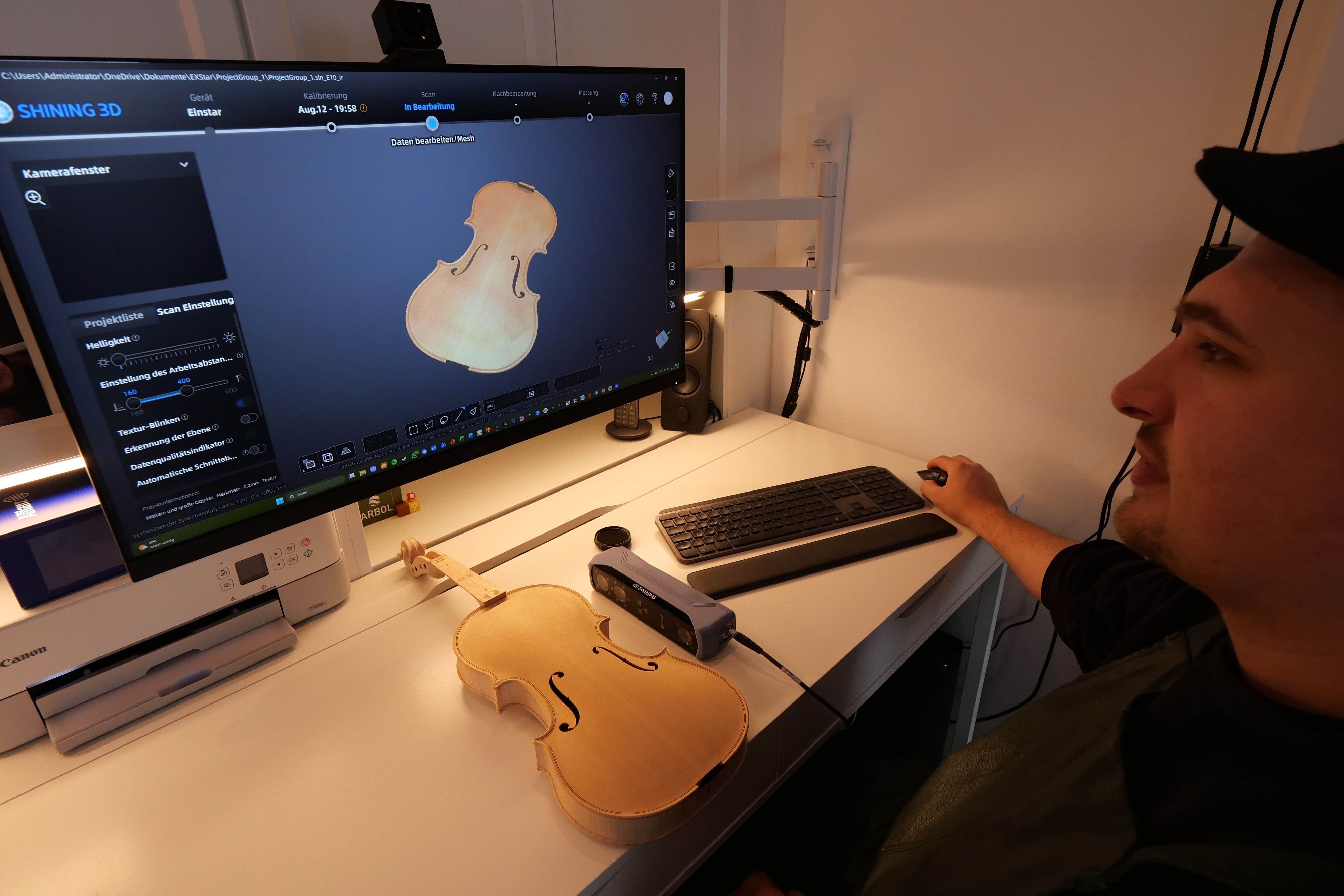 Ein Mann mit Kappe sitzt vor einem Monitor, auf dem der Korpus einer Geige zu sehen ist. Eine echte Geige liegt vor ihm auf dem Schreibtisch.
