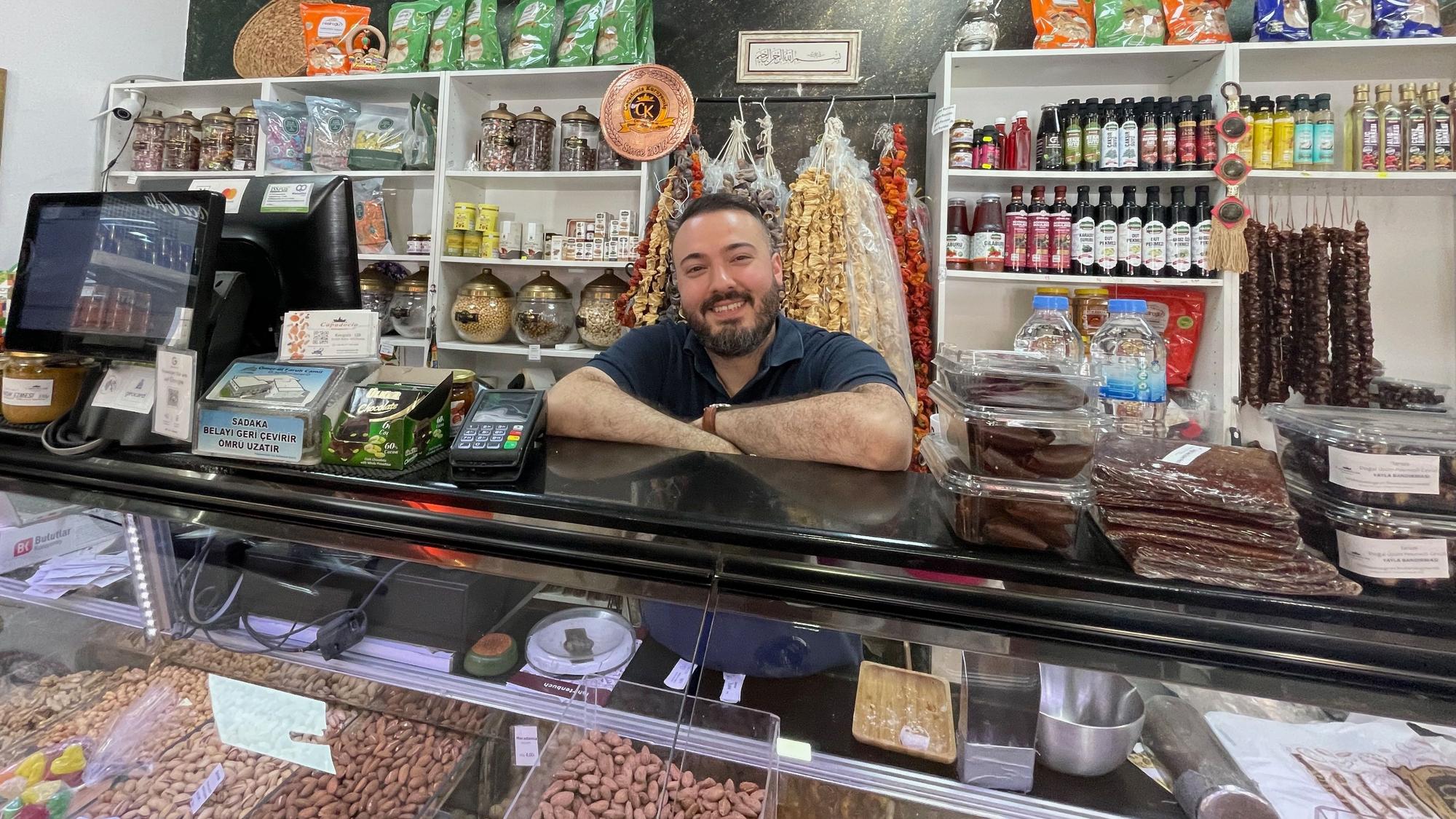 Ein junger Mann mit dunklem Haar und Bart steht hinter einer Frischetheke in einem Laden.