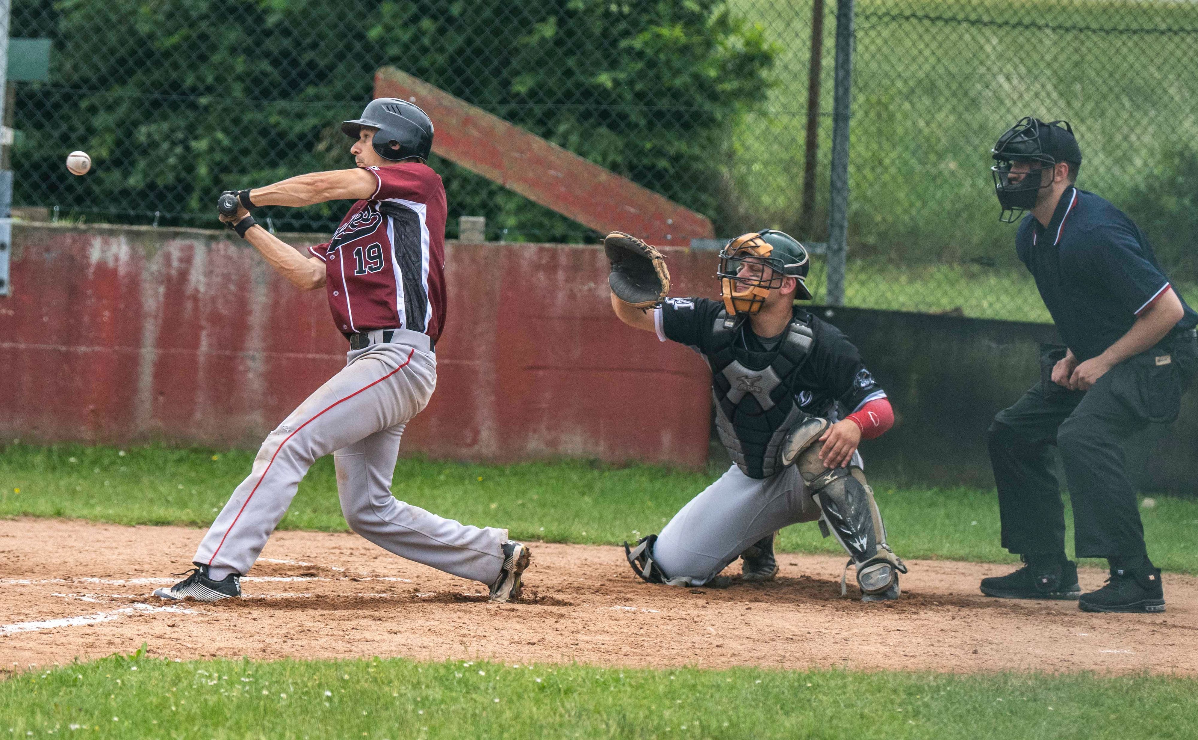 Ein Baseballspieler beim Schlag. Die Muskeln sind angespannt. Hinter ihm der Catcher und der Umpire.