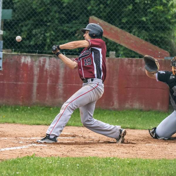 Ein Baseballspieler beim Schlag. Die Muskeln sind angespannt. Hinter ihm der Catcher und der Umpire.
