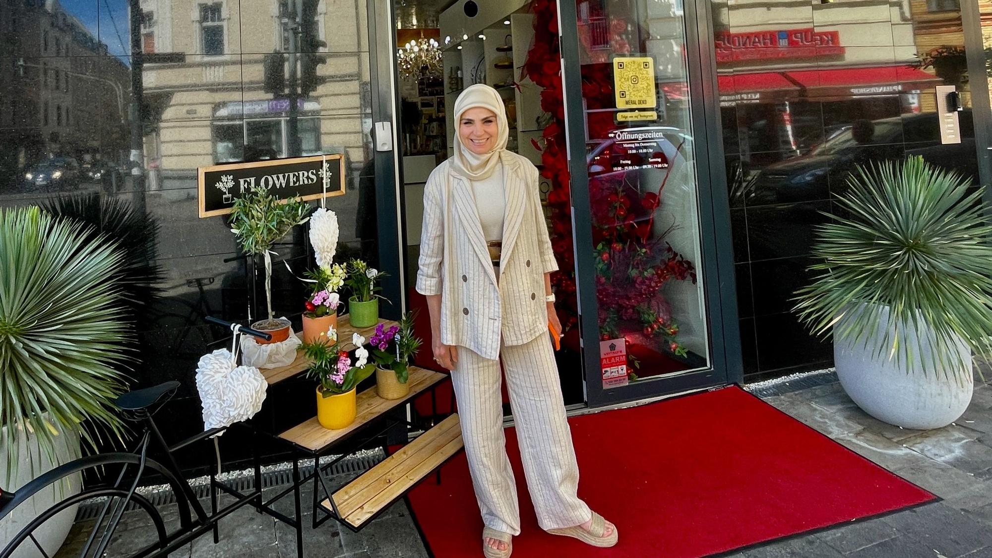 Eine Frau in hellem Blazer, heller Hose und hellem Kopftuch steht vor dem Schaufenster ihres Ladens.
