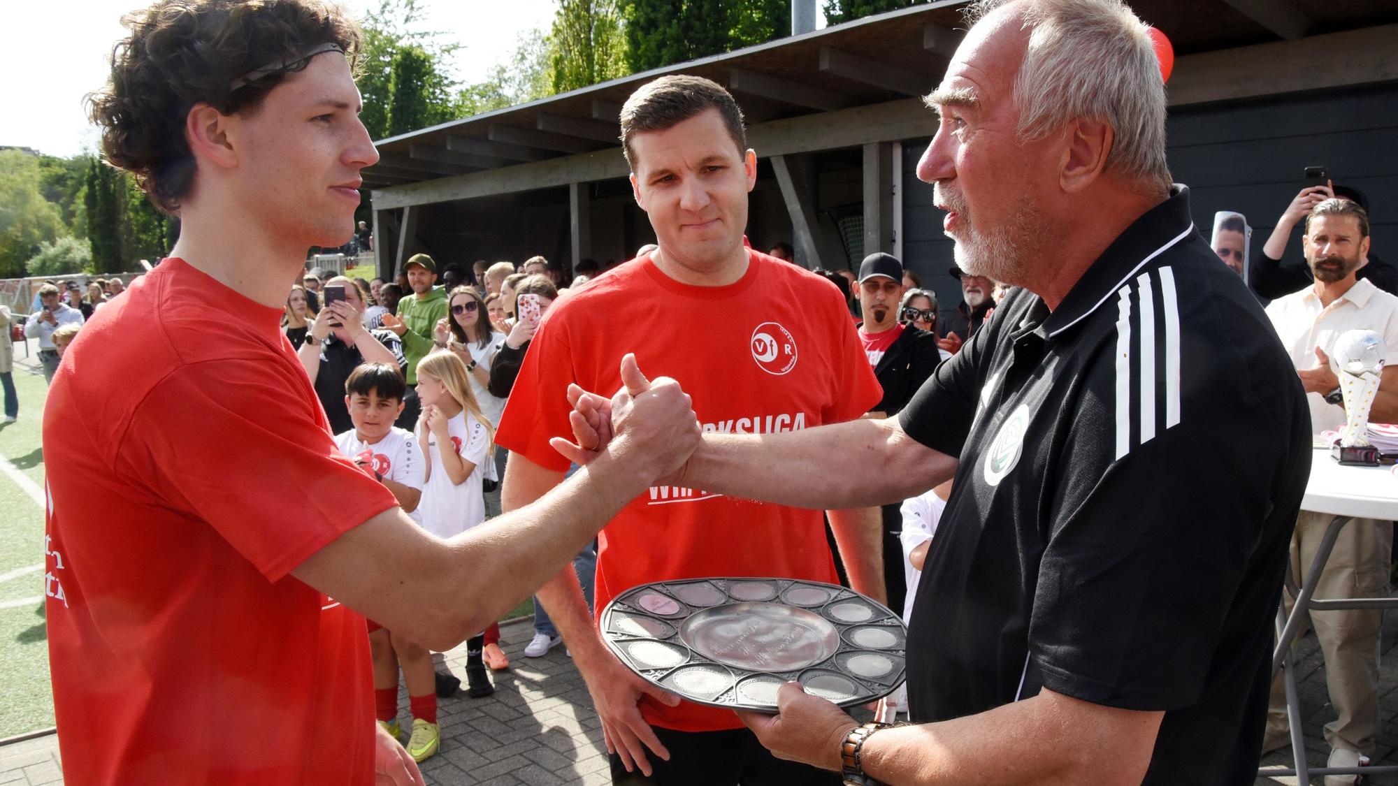 Zu sehen ist die Übergabe einer Meisterschale durch einen Sportfunktionär an zwei Fußballspieler.