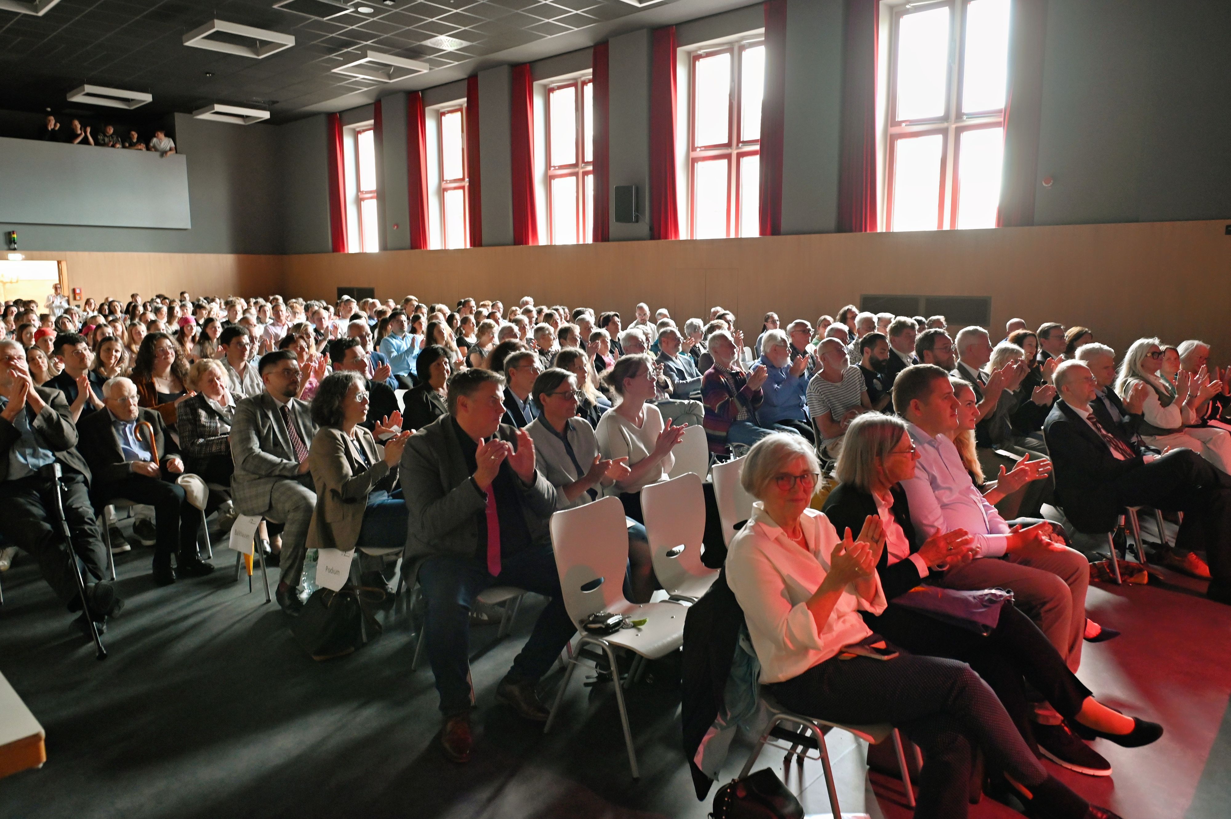 Zahlreiche Menschen sitzen auf Stühlen in der Aula der Gesamtschule Euskirchen und applaudieren.