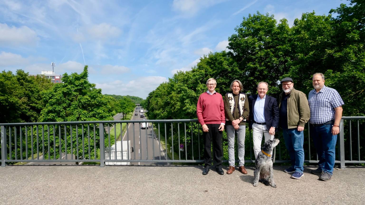 Fünf Männer und ein Hund stehen auf einer Brücke über einer vielbefahrenen, mehrspurigen Straße.