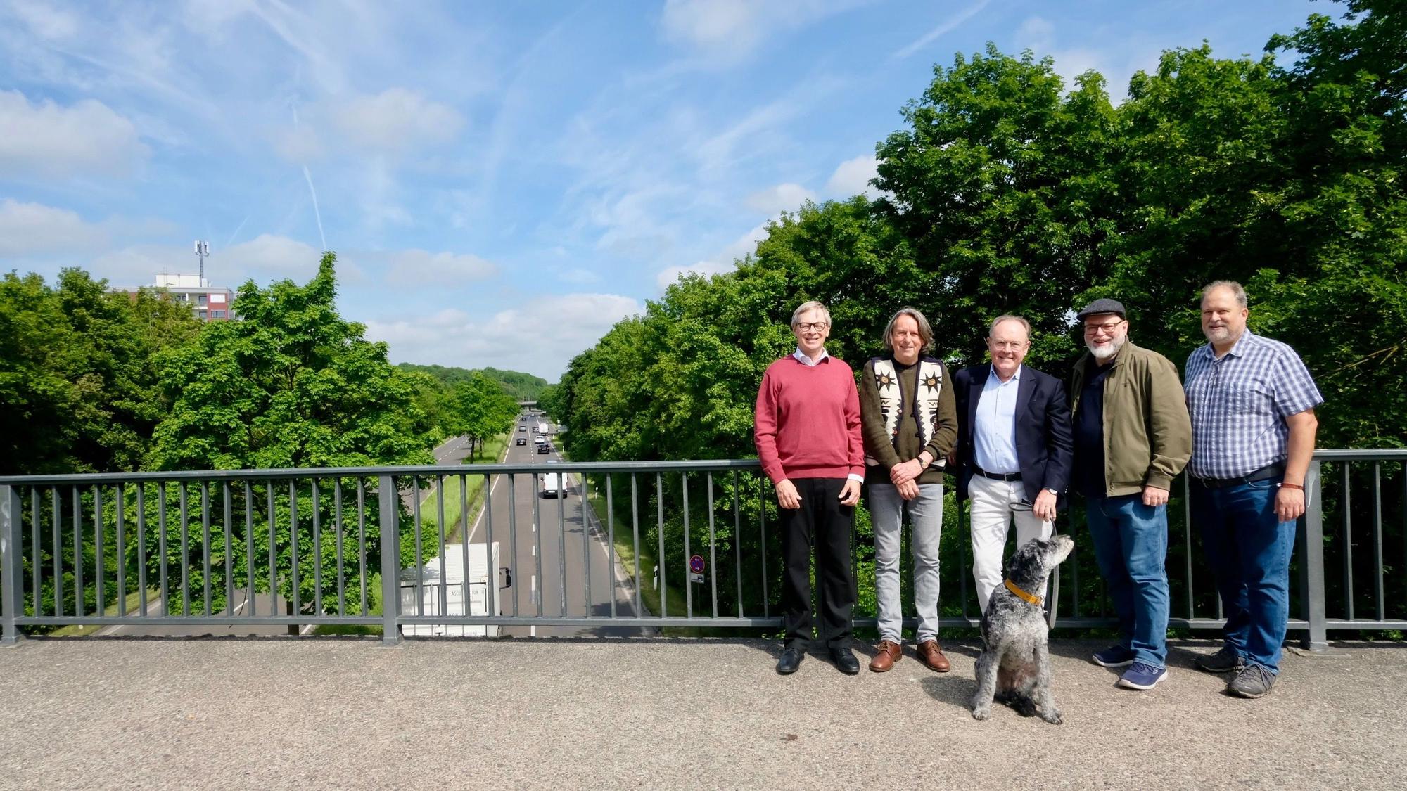 Fünf Männer und ein Hund stehen auf einer Brücke über einer vielbefahrenen, mehrspurigen Straße.