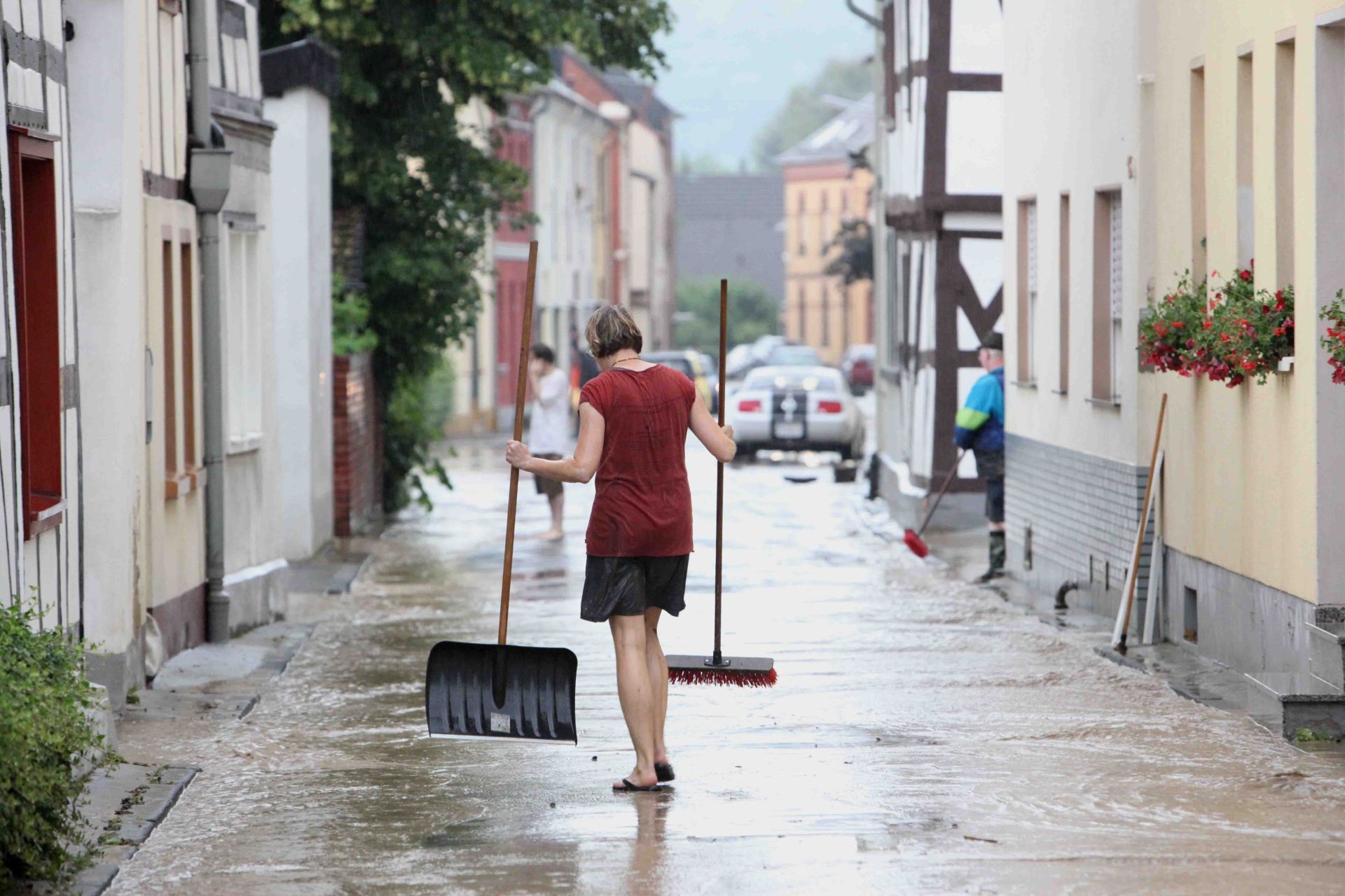 Eine Frau mit Besen und Schaufel auf einer von braunem Wasser überfluteten Straße.