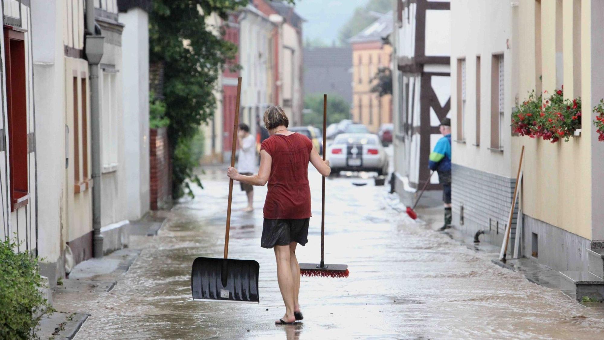 Eine Frau mit Besen und Schaufel auf einer von braunem Wasser überfluteten Straße.