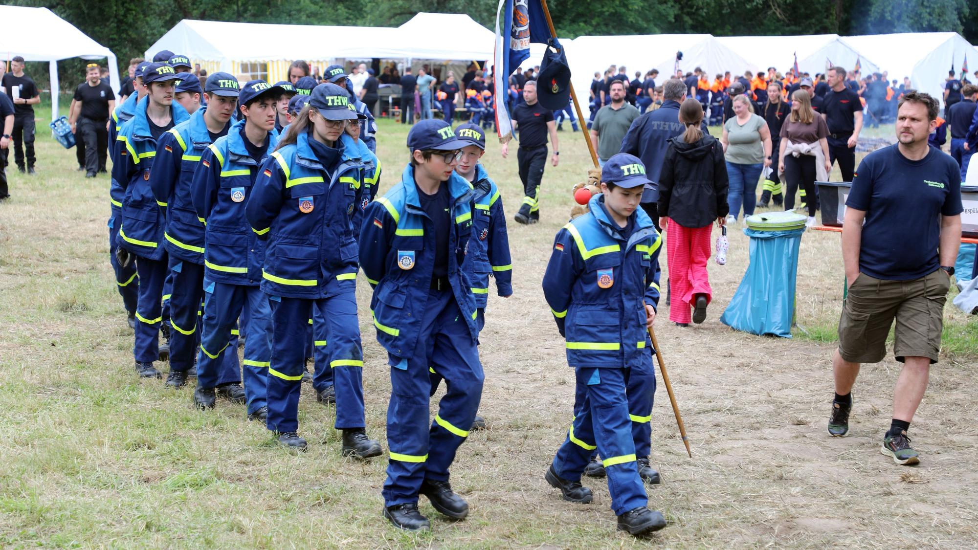 Eine THW-Jugendgruppe marschiert mit Wimpel über den Zeltplatz des Kreisjugendfeuerwehrzeltlagers 2026 in Bergisch Gladbach-Paffrath.