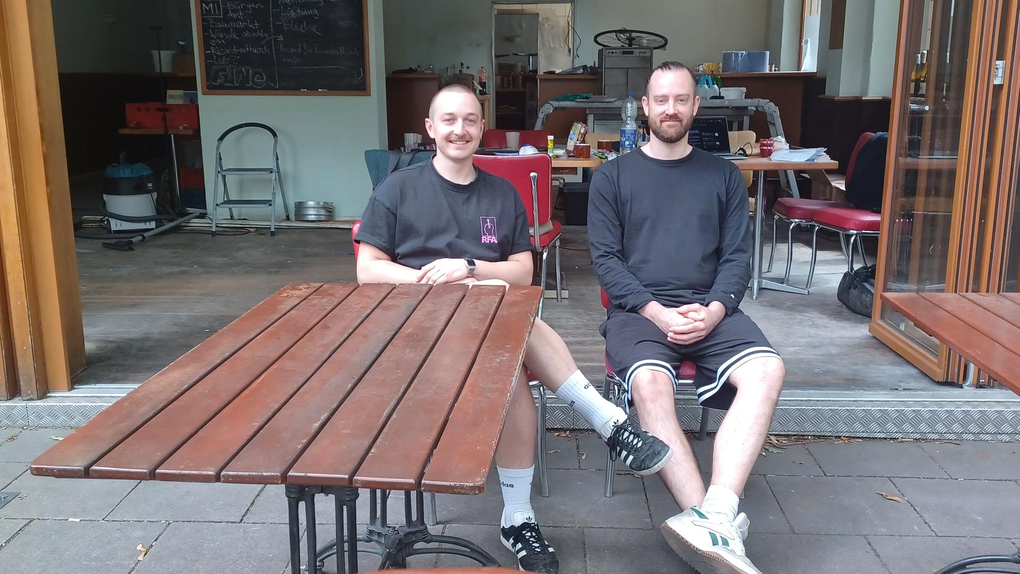 Zwei junge Männer sitzen auf einer Terrasse vor einem Café, das gerade umgebaut wird.