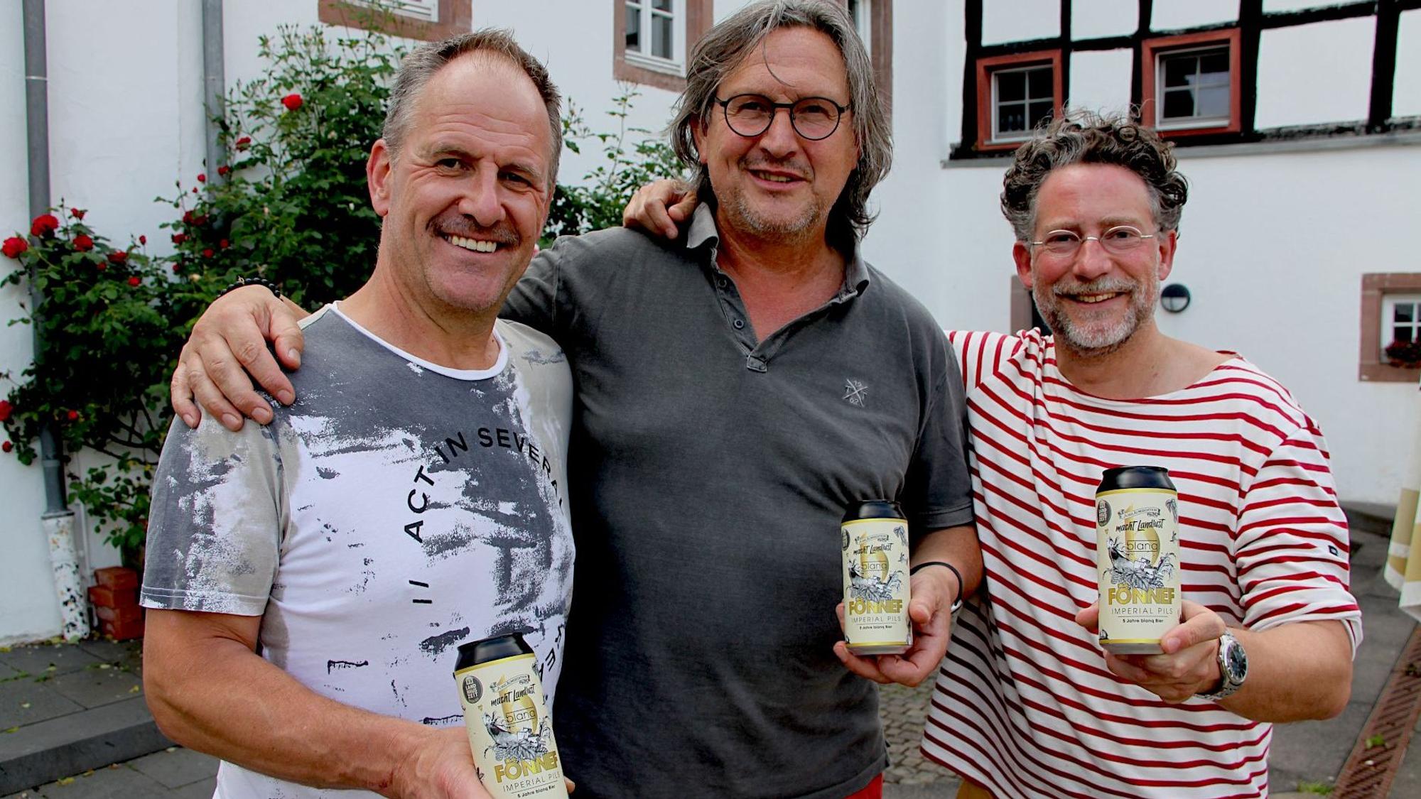 Drei Männer stehen Arm in Arm vor einem Fachwerkhaus in Blankenheim. Jeder hält eine Dose Bier in der Hand.