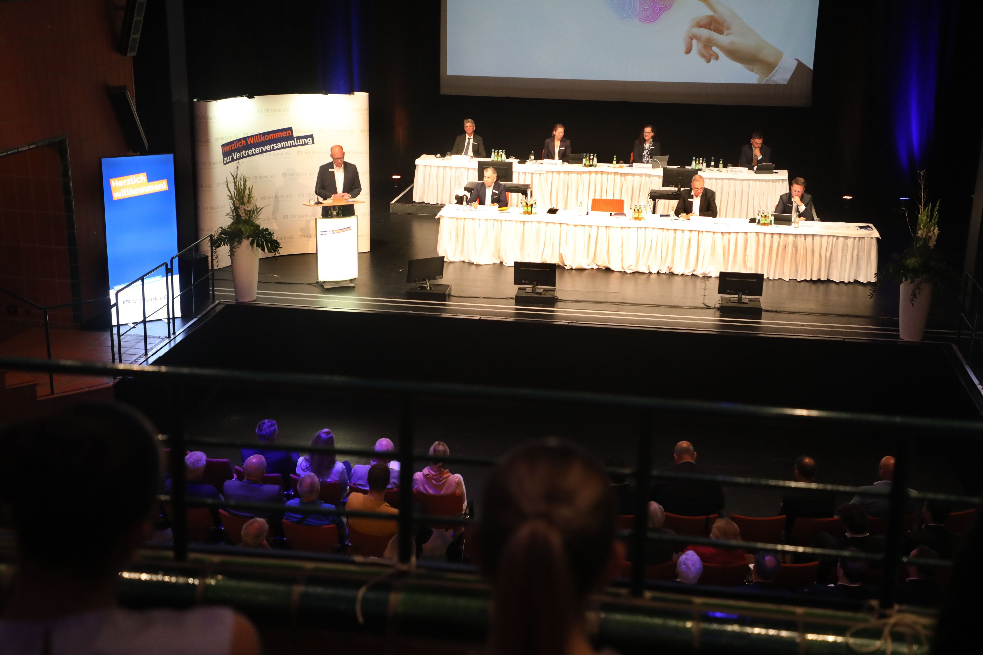 Auf einer Bühne stehen zwei lange Tischen an denen Menschen sitzen und ein Mann steht an einem Rednerpult.