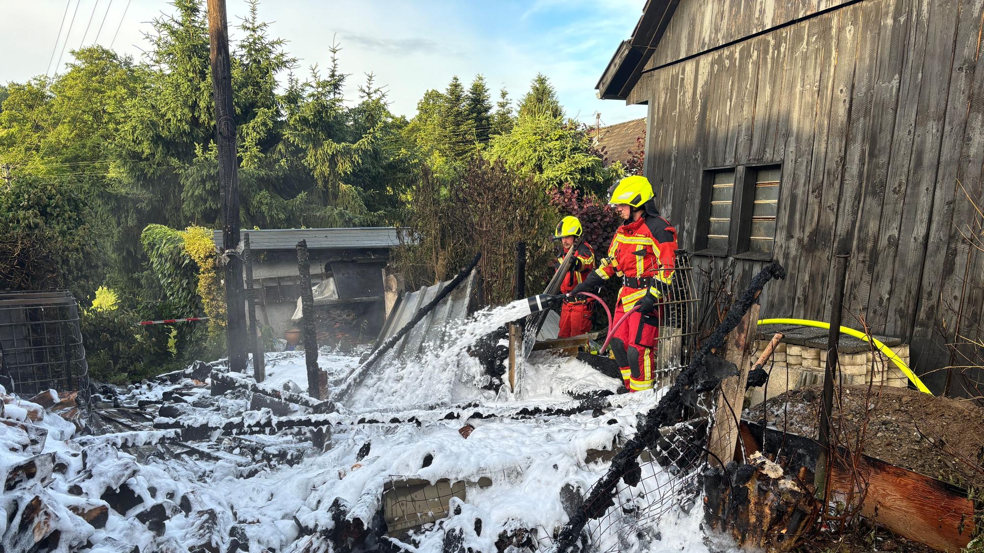 Das Foto zeigt die Reste eines abgebrannten Schuppens und zwei Feuerwehrleute