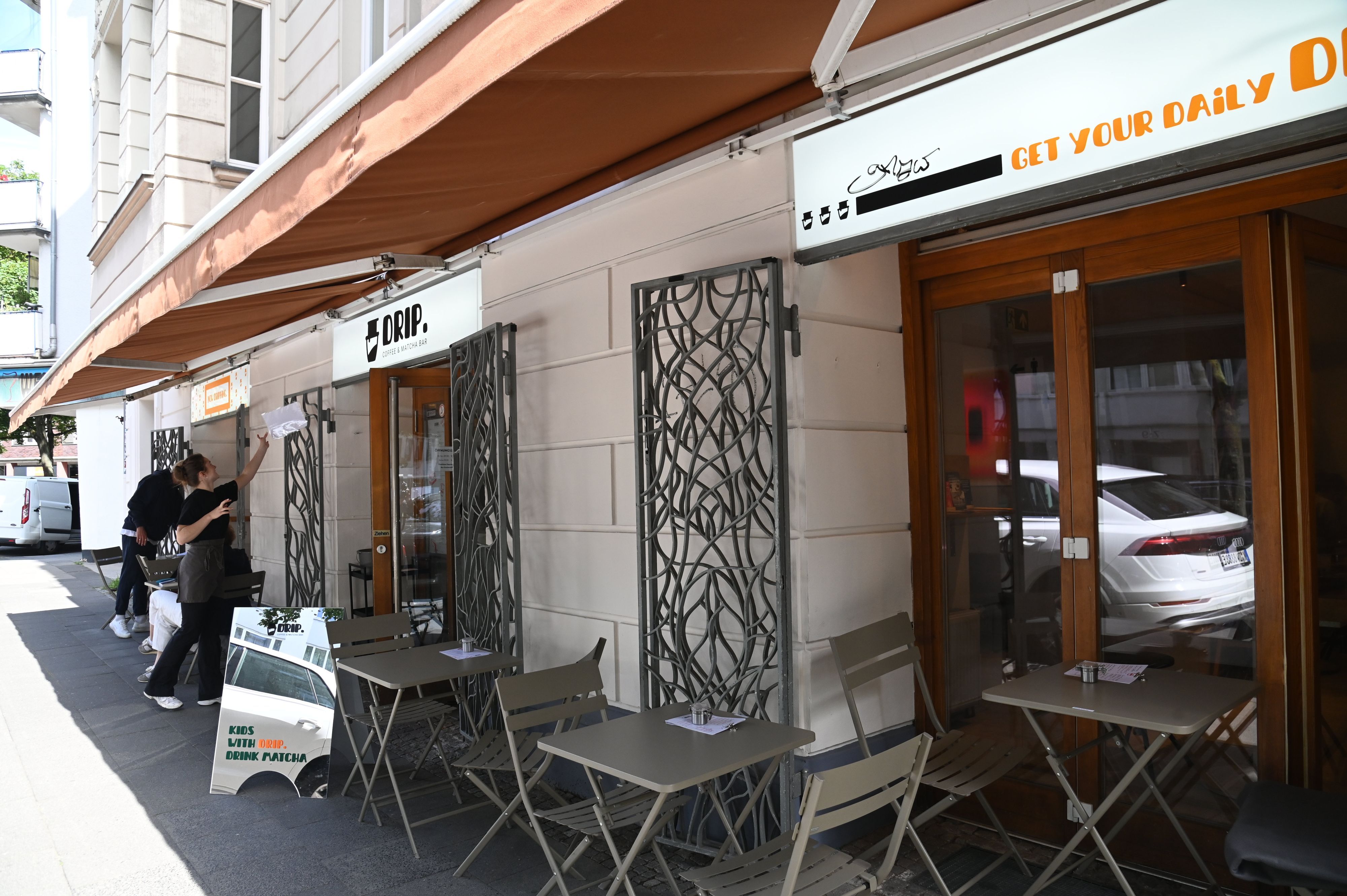 Das Drip-Coffee von außen. Vor dem Laden stehen Tische und Stühle zum Draußensitzen unter einer terrakottafarbenen Markise.