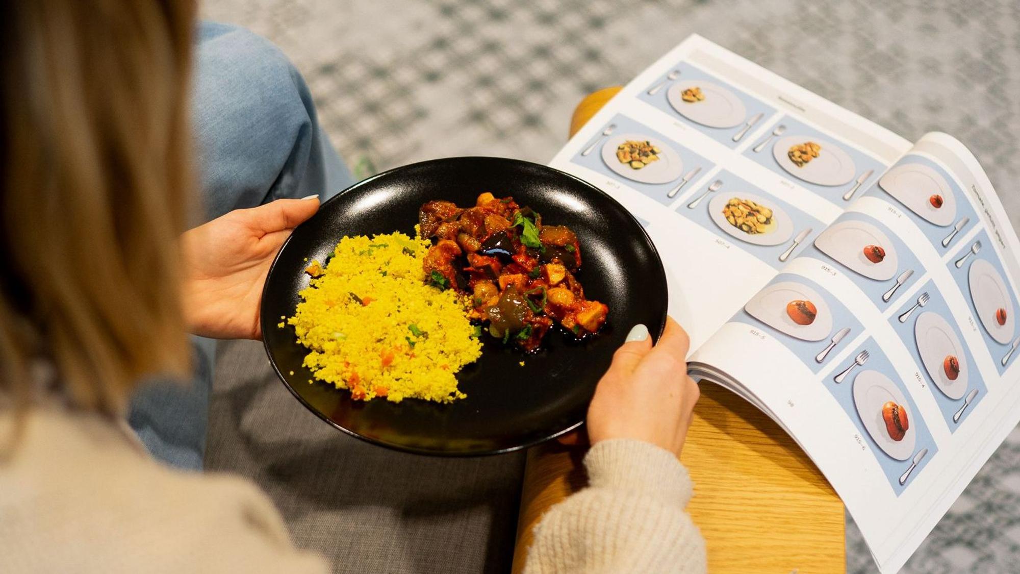 Das Foto zeigt eine Frau von hinten, die einen Teller mit einer Speise in den Händen hält, davor ein Buch mit Abbildungen von Speise-Portionsgrößen.