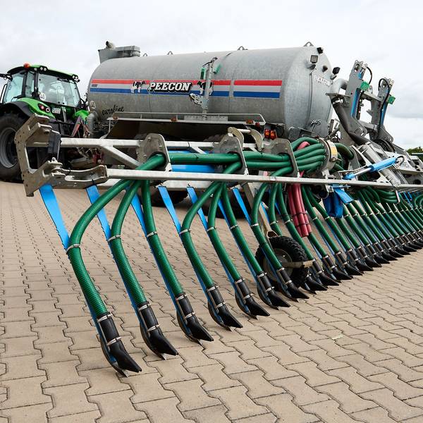 Ein Traktor steht auf einer gepflasterten Fläche. Er zieht ein großes Fass mit einer Vorrichtung, die Gülle dicht am Boden ausbringt.