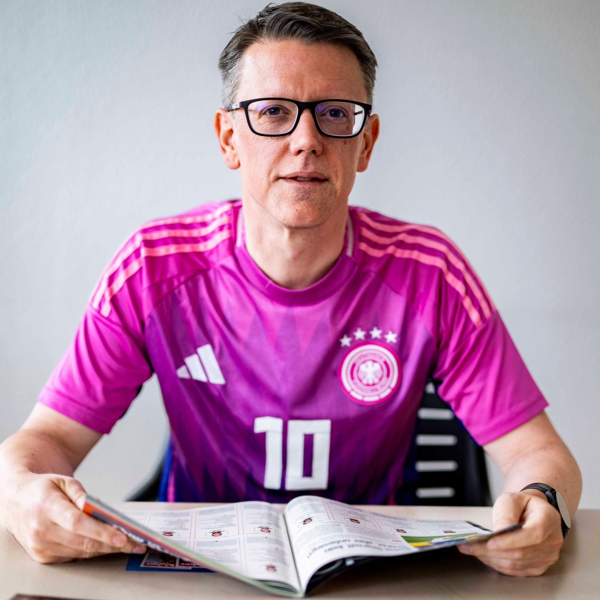 Thomas Schmitz sitzt im deutschen Auswärtstrikot der WM 2022 am Tisch und blättert im Kicker-Sonderheft.
