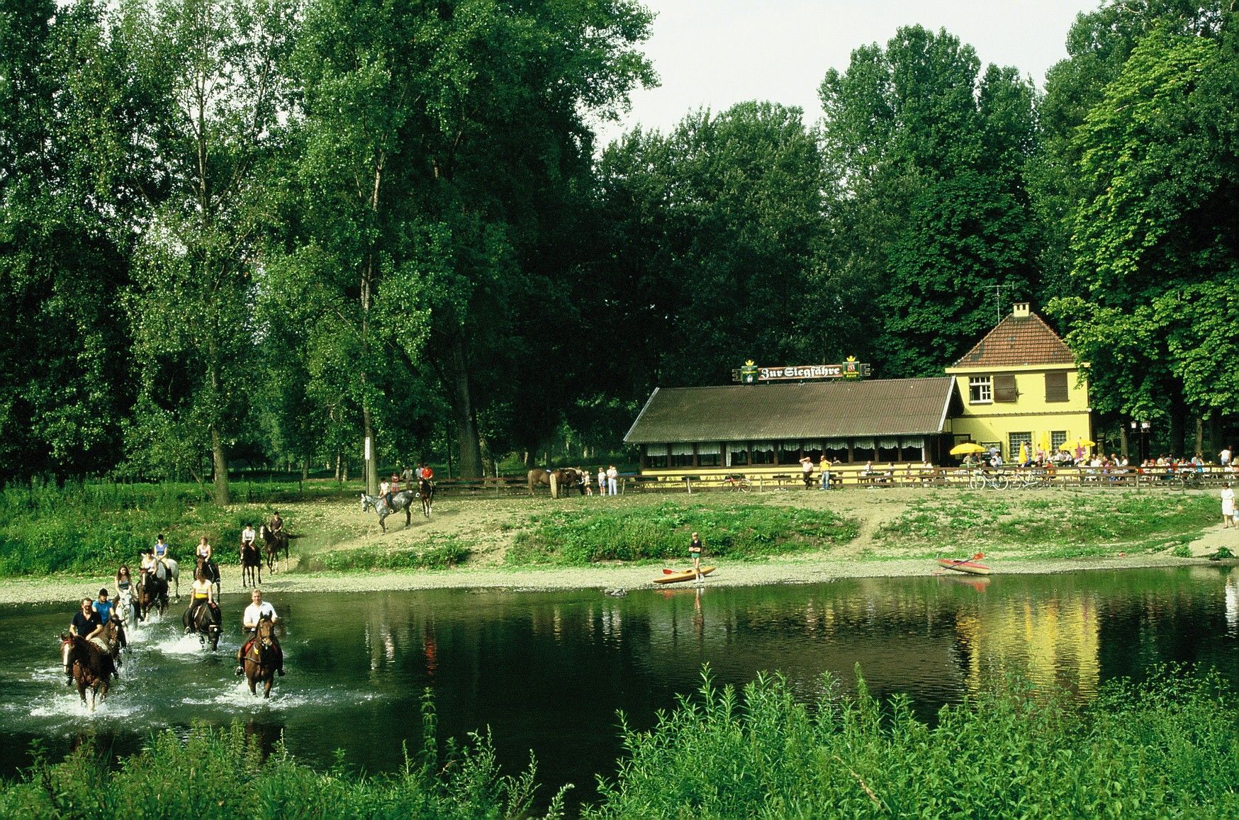 Die Aufnahme zeigt eine Gruppe, die mit Pferden die Sieg überquert. Im Hintergrund ist das Restaurant „Zur Siegfähre“ zu sehen.
