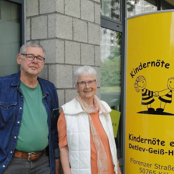Zwei Männer und eine Frau stehen links und rechts von einer gelben Säule, auf der Kindernöte e.V., steht.